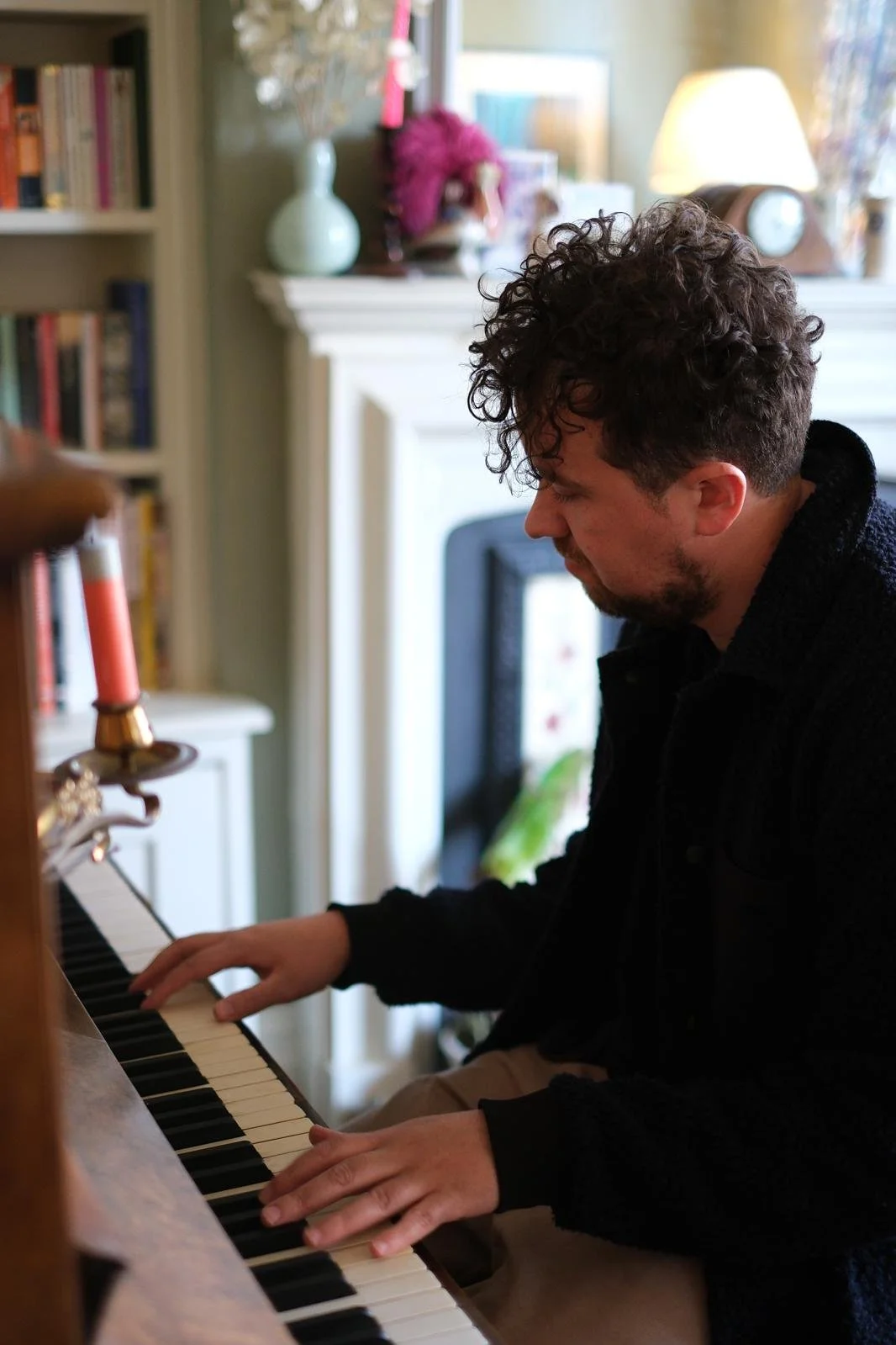A man with curly hair and a beard playing a piano in a cozy, decorated room, with a bookshelf, lamp, and decorative items in the background.
