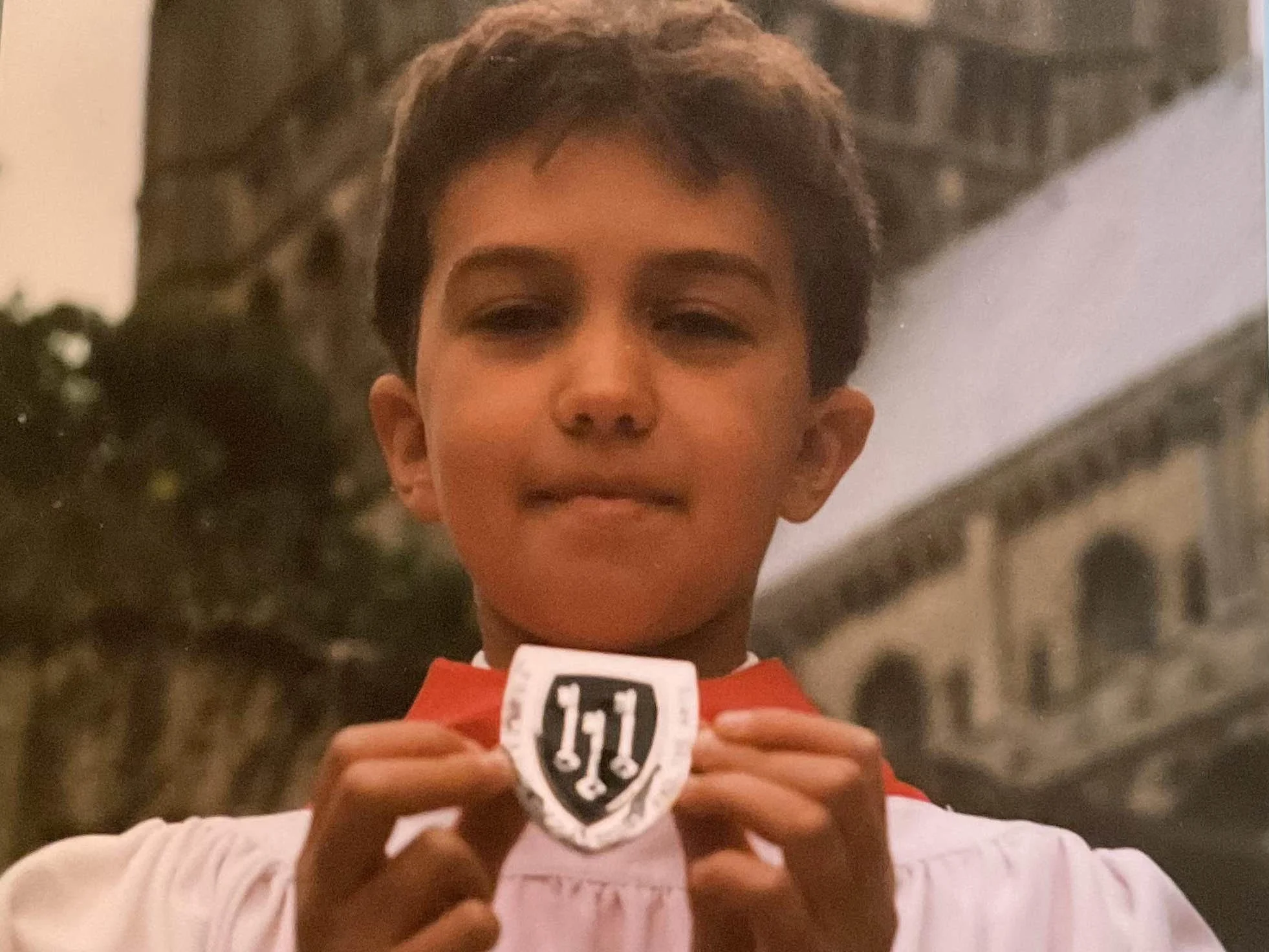 A young boy chorister with short dark hair is wearing red and white robes and holding a medal up to the camera with three keys on it