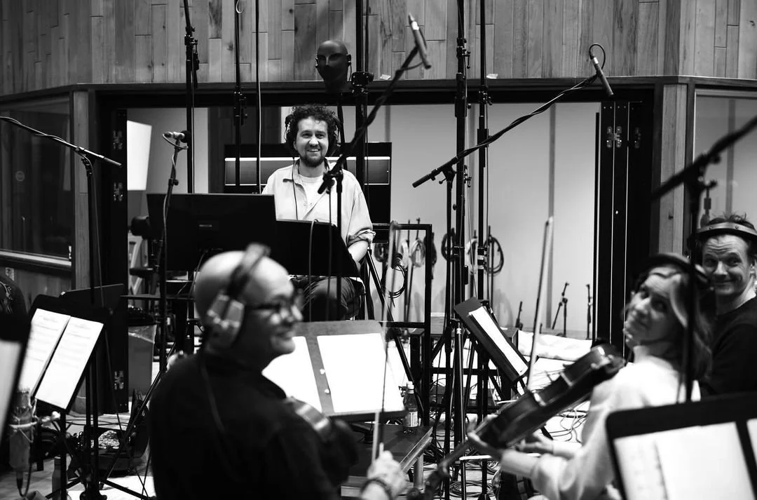 Black and white photo of musicians in a recording studio, with a man smiling at the microphone. The studio has wooden walls, microphones hanging from above, and music stands with sheet music.