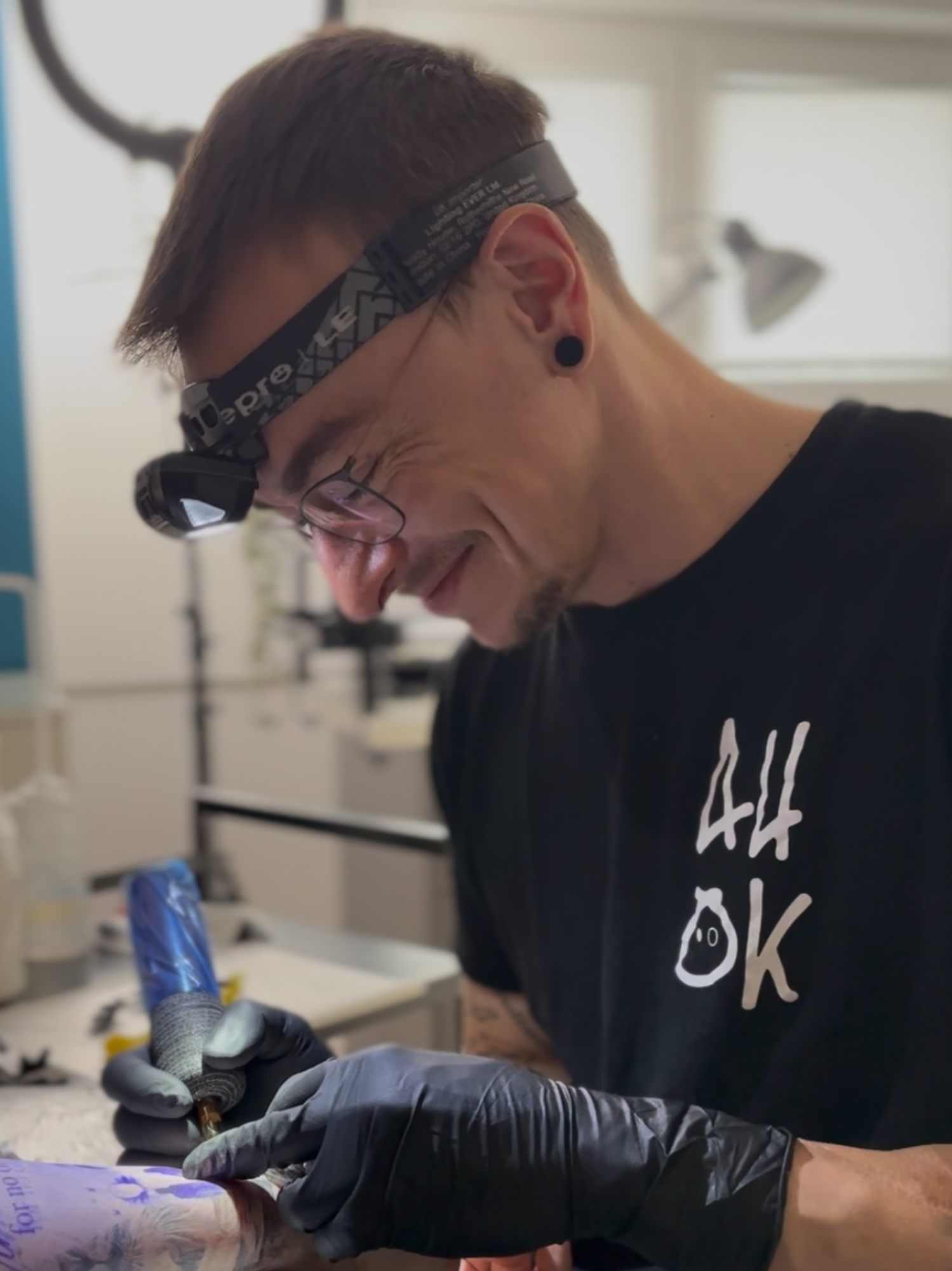 Mann mit Brille, Headlamp und E-Ohrring, arbeitet an einem Tattoo an einem Arm, trägt schwarze Handschuhe, im Hintergrund ein Behandlungsraum.