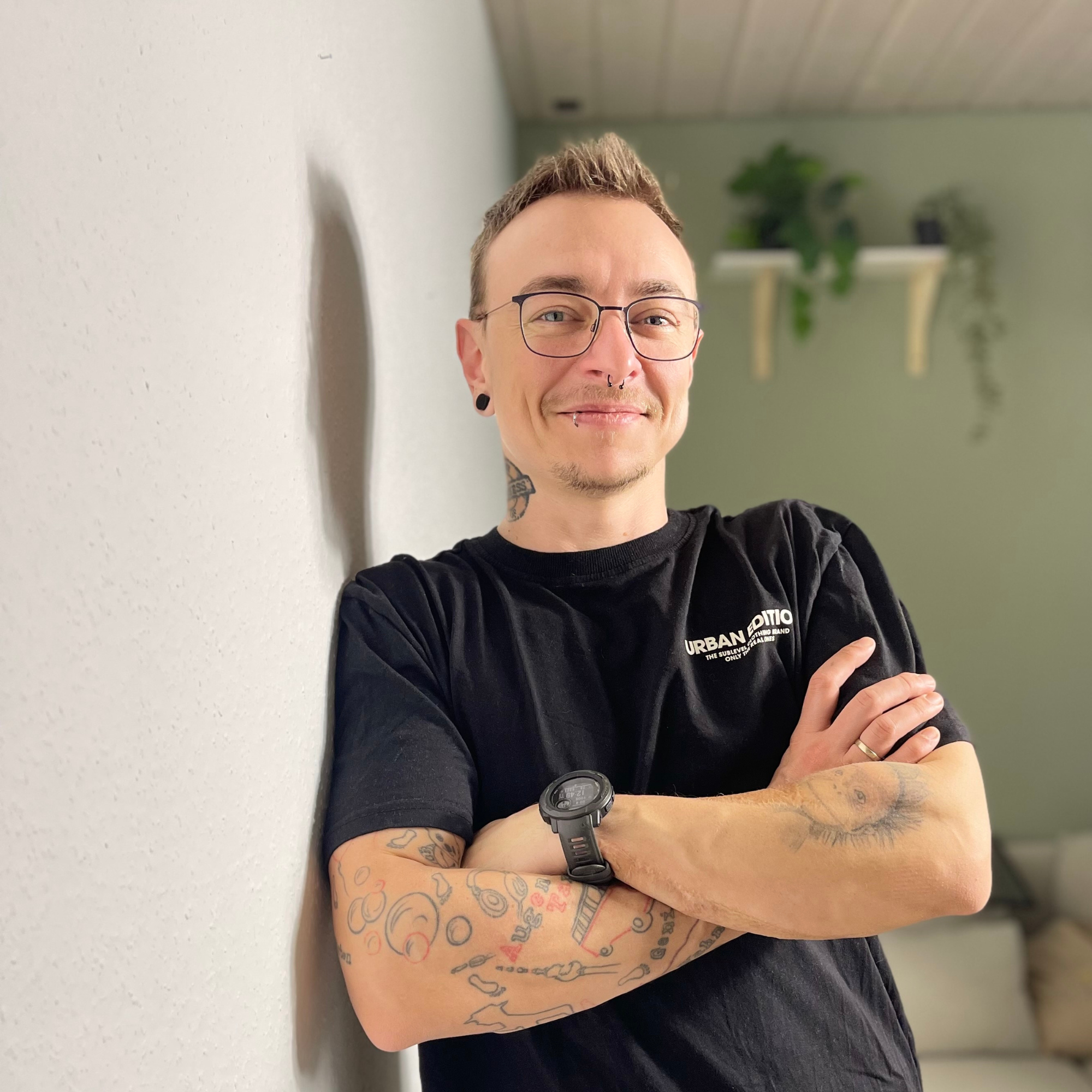 Junge Frau mit Tattoos, Brille, schwarzen T-Shirt mit Aufdruck, Armbanduhr, steht an Wand, lächelt, mit Arm vor Brust. Wohnzimmer mit Pflanzen im Hintergrund.