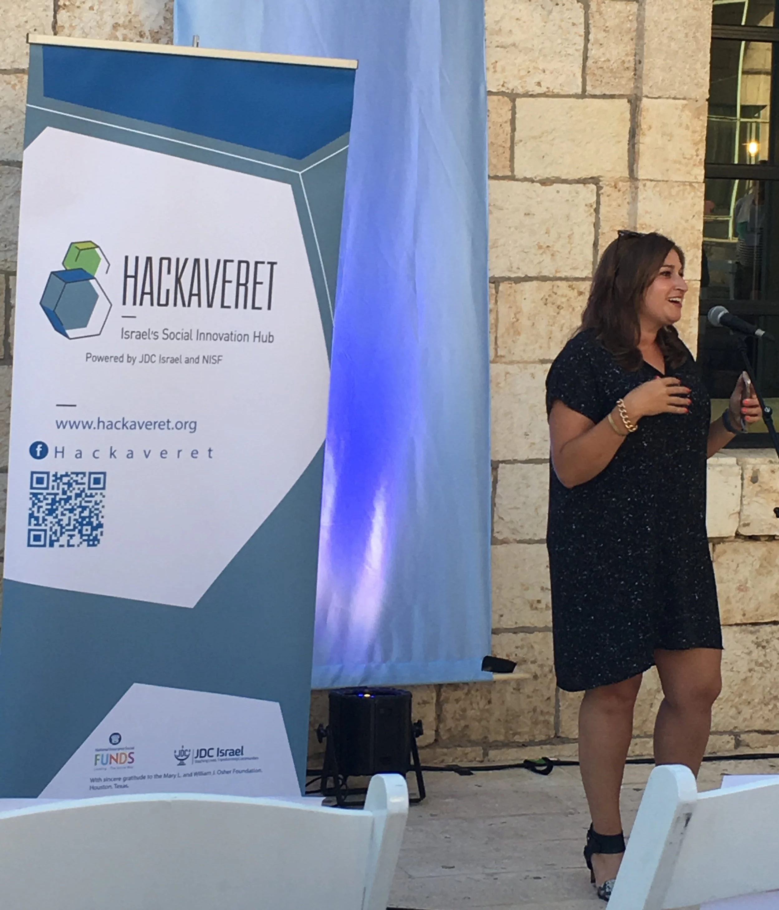 A woman speaking at a conference during an event named Hackaveret, which is Israel's Social Innovation Hub. The woman is standing next to a banner with the Hackaveret logo and website, wearing a black dress with floral patterns, and holding a phone. The setting appears to be outdoors with a stone wall in the background.