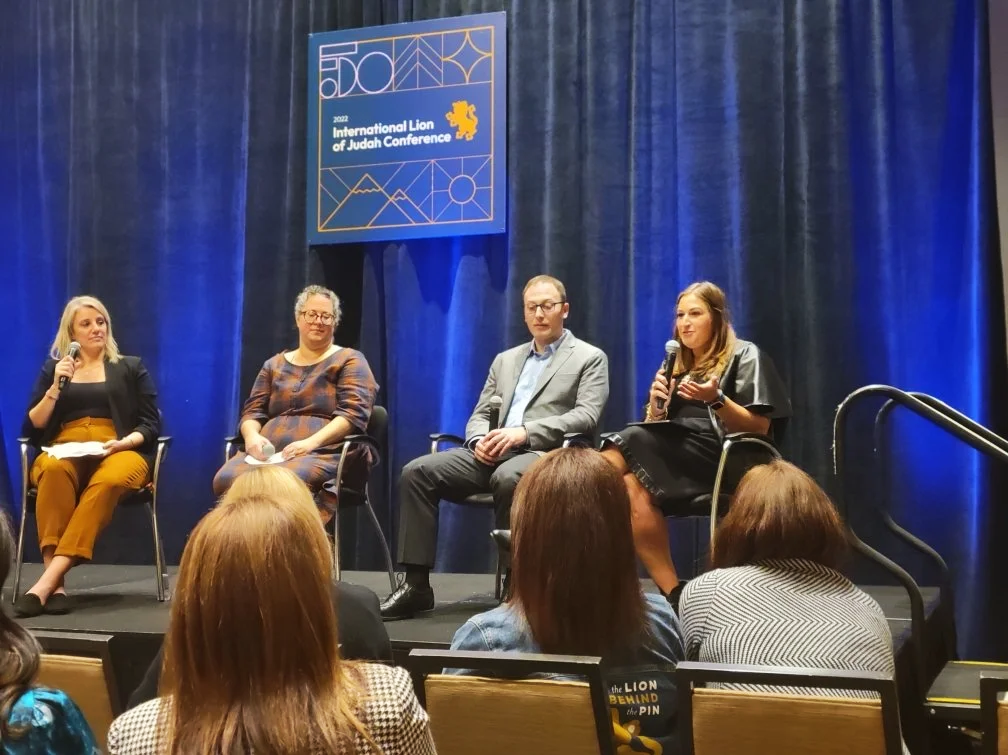 Panel discussion at the 2022 International Lion of Judah Conference, with four women and one man seated on stage, speaking into microphones, with an audience in front and a blue curtain backdrop.
