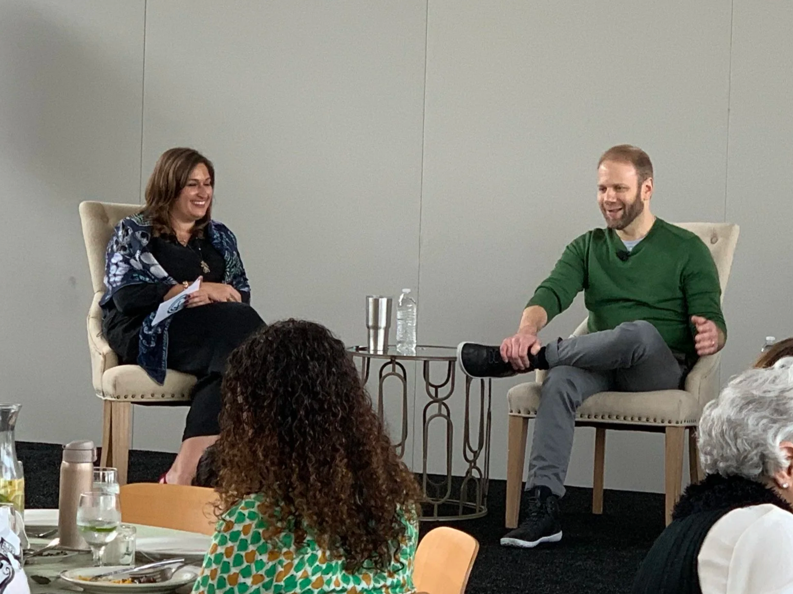 A woman and a man sit on stage during a discussion, both smiling. The woman has shoulder-length brown hair and is wearing a dark outfit with a patterned shawl. The man has a beard, short hair, and is wearing a green sweater and gray pants. There is a small table between them with water bottles, a cup, and a microphone. Audience members are visible in the foreground.