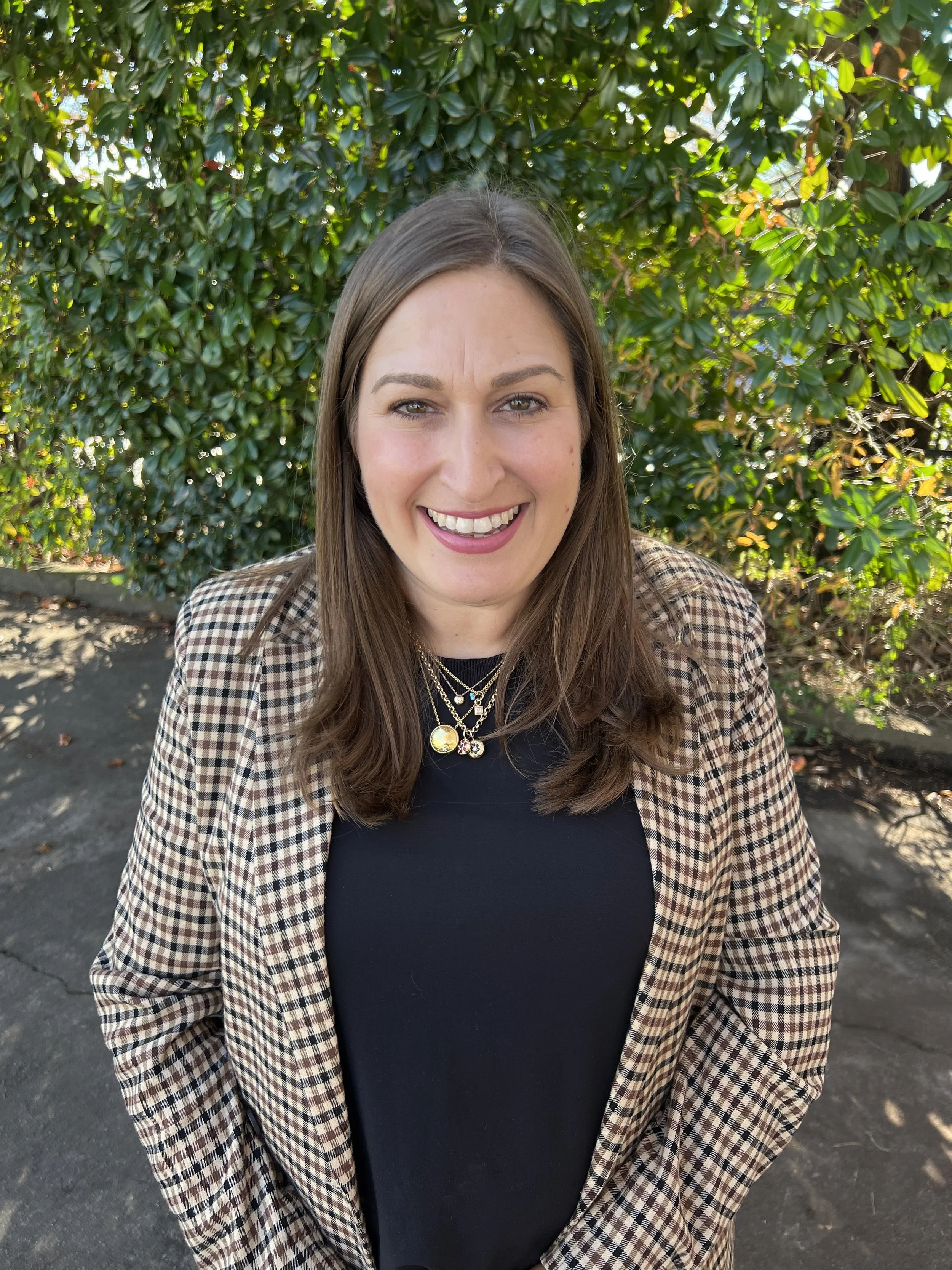 A woman smiling outdoors, with long brown hair, wearing a plaid blazer and layered necklaces, standing in front of dense green shrubbery.