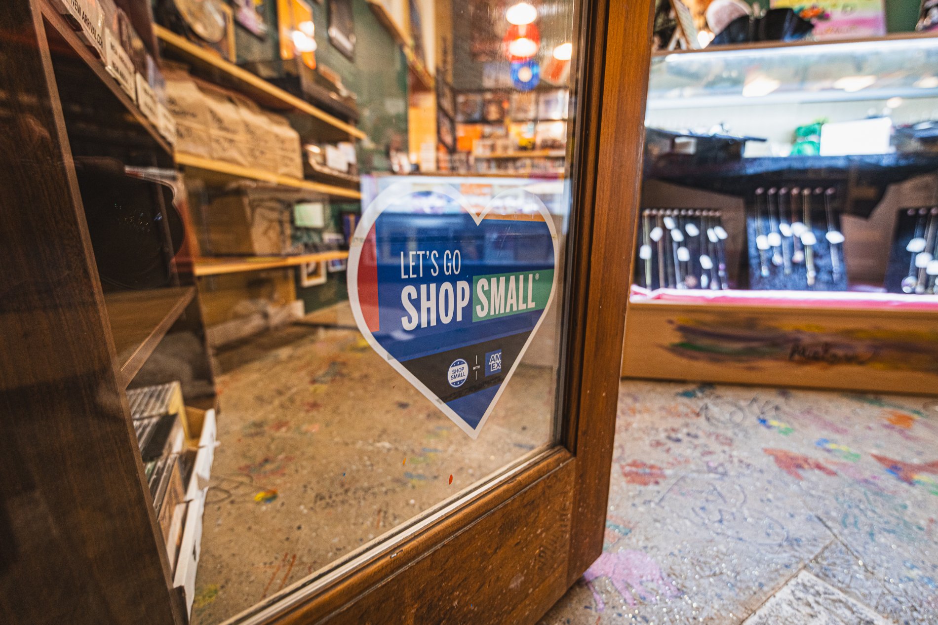 Close-up of a glass door with a sticker promoting small shops, reading "Let's go shop small," inside a store with shelves filled with various items, and a colorful, slightly blurred background.