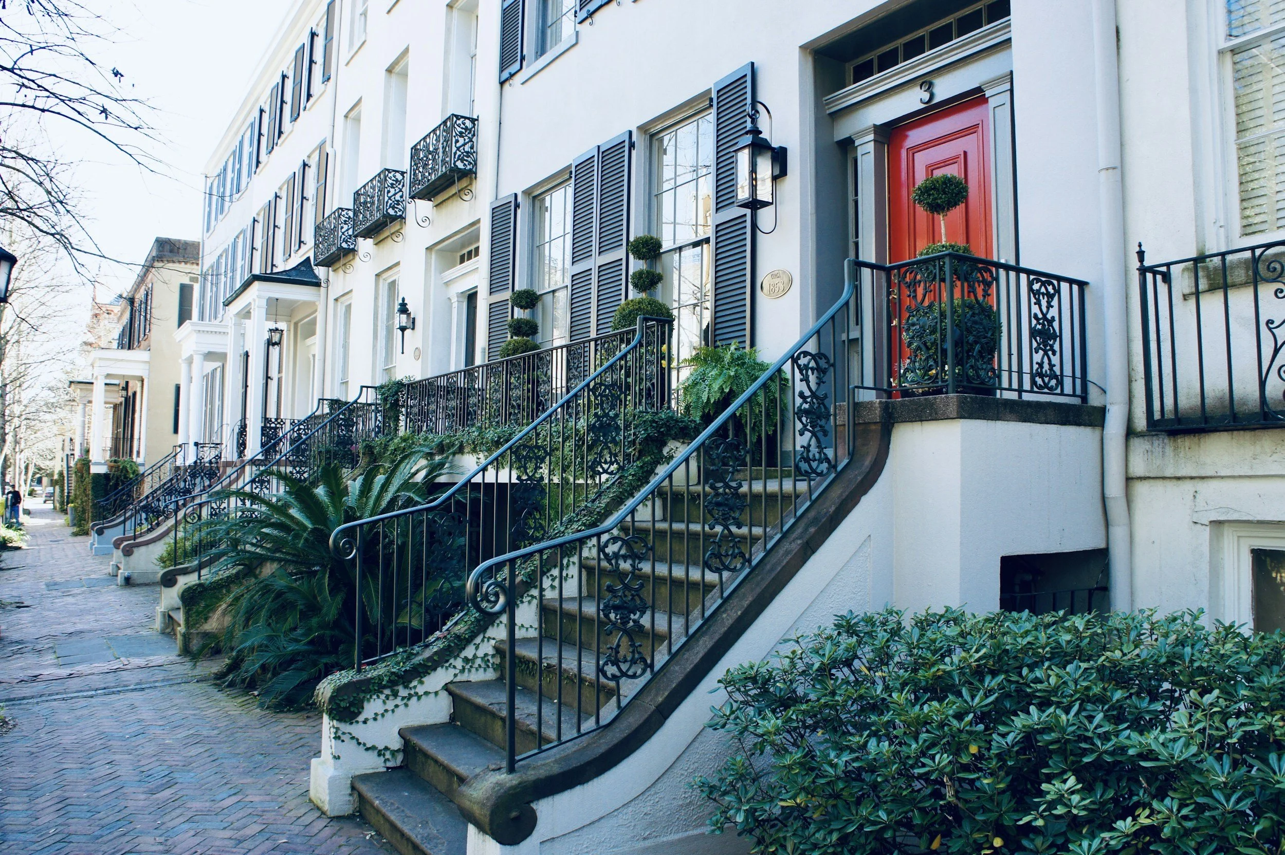 Exterior view of a row of modern townhouses with stairs leading to the front doors, featuring black wrought iron railings, potted plants, and a red door, on a cobblestone street.