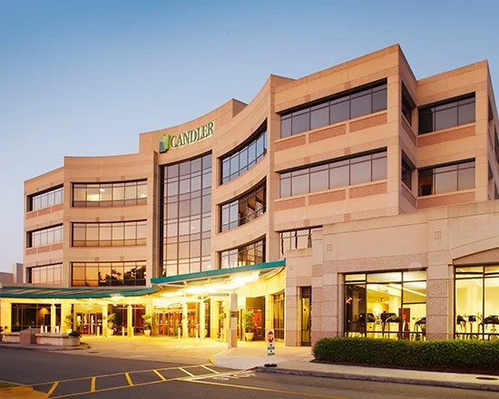 Modern multi-story building with the sign 'Candler' on the top, large glass windows, and a brightly lit entrance at dusk, with a parking lot in front.