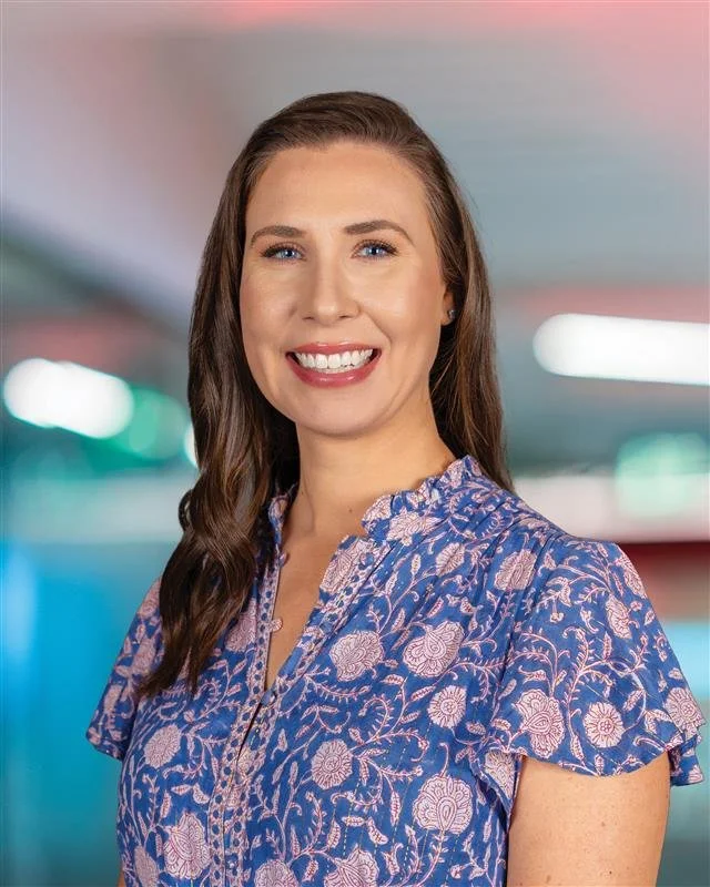 A smiling woman with long brown hair, wearing a blue floral blouse, in an indoor setting with colorful, blurred background lights.