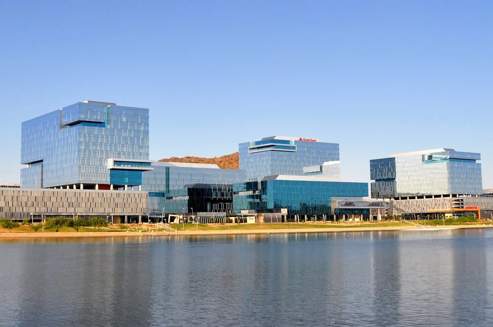 Modern glass building complex by a body of water with mountains in the background, clear blue sky.