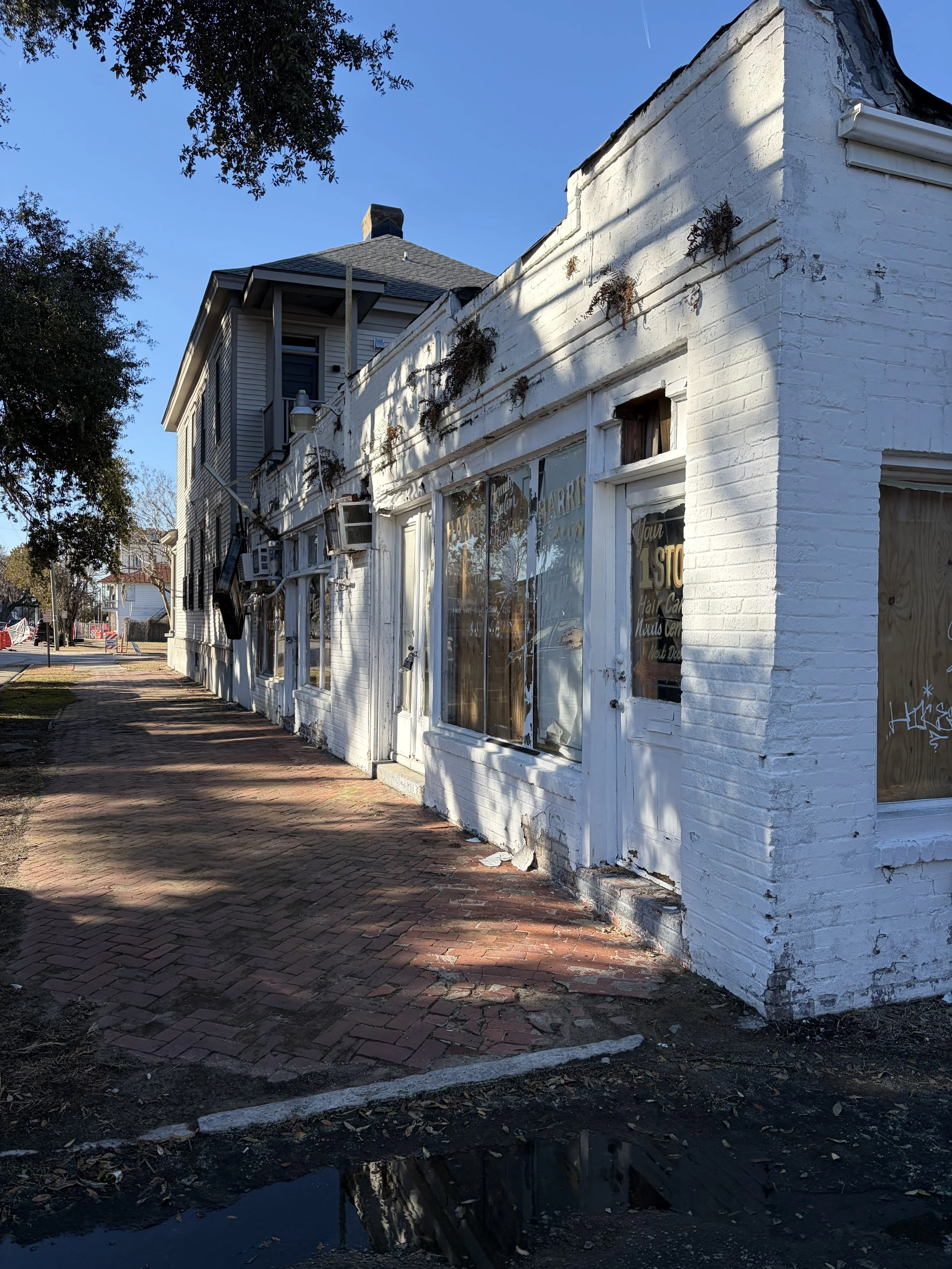 Old white brick building with boarded-up windows and door, some windows have graffiti, injury to the paint and structure at the bottom, outside sidewalk with brick paving, trees casting shadows on the building, clear blue sky.