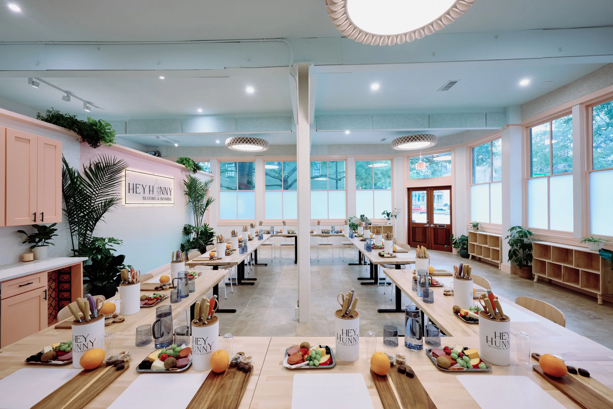 A bright, spacious room set up for a cooking class or gathering, with tables arranged in a U-shape, each with ingredients, utensils, and glasses. The room has large windows, plants, and modern lighting.