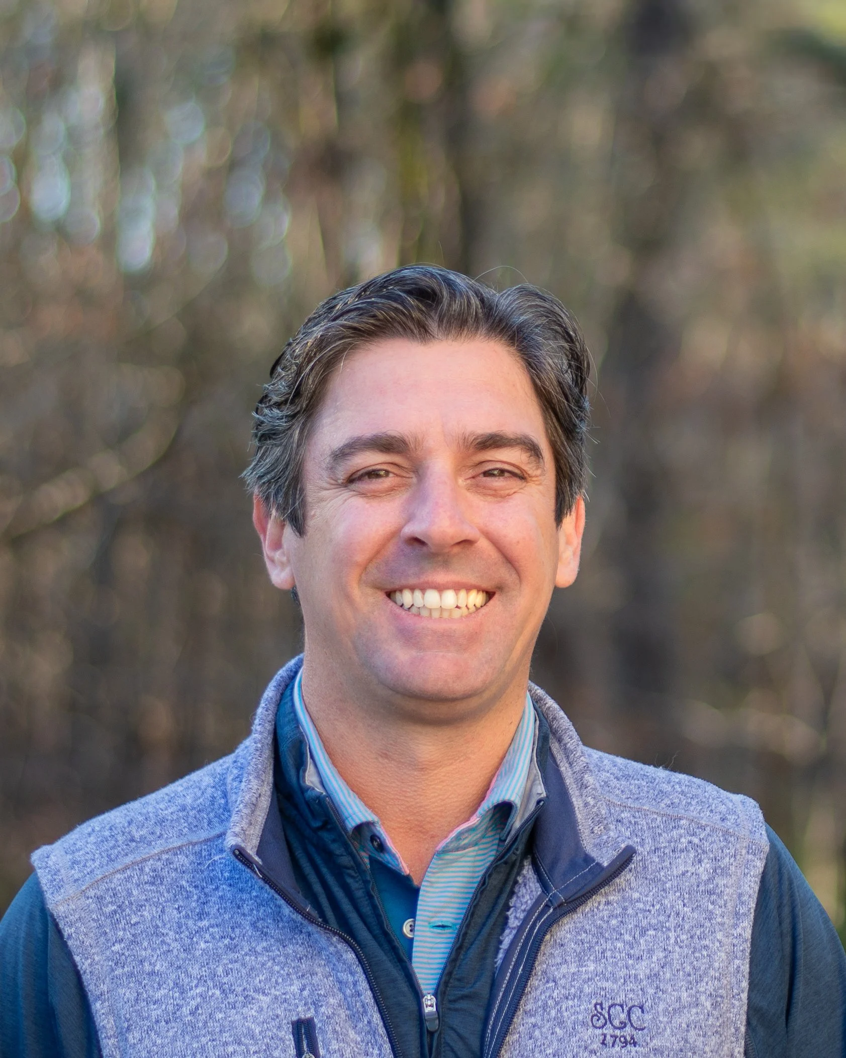 A smiling man with dark hair standing outdoors in front of a wooded area.