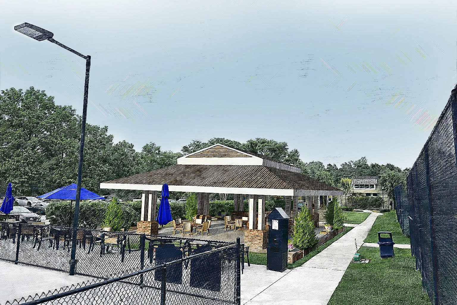 An outdoor seating area with tables and chairs under a large, multi-sided pavilion with a brown shingled roof. Blue umbrellas are open over some tables. There is a paved pathway, a trash can, and a black fence surrounding the seating area. Parking lot with cars and greenery in the background.