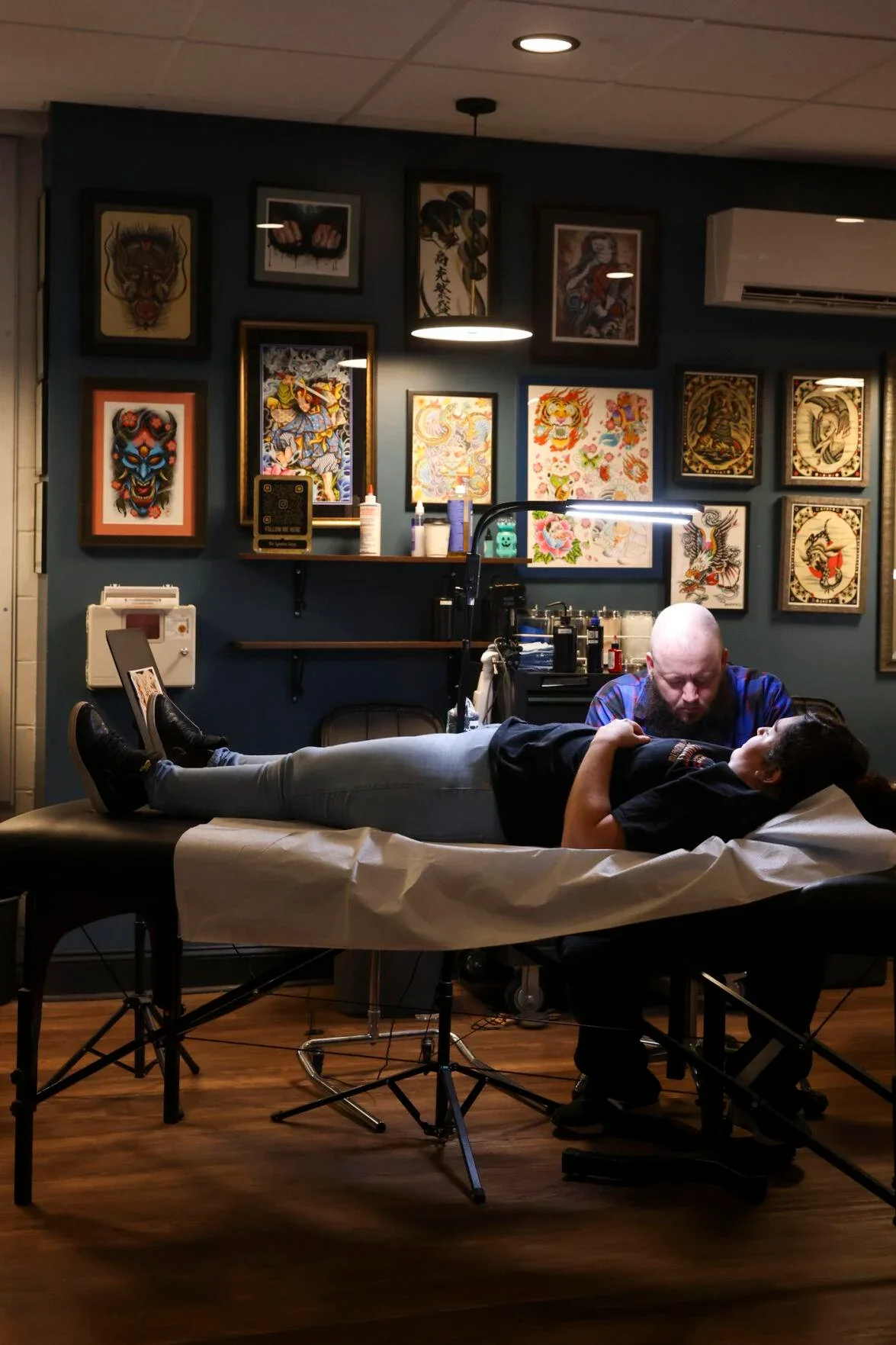 A person lying on a tattoo artist's table getting a tattoo, while the tattoo artist works on their side. The room features framed tattoo artwork on the wall.