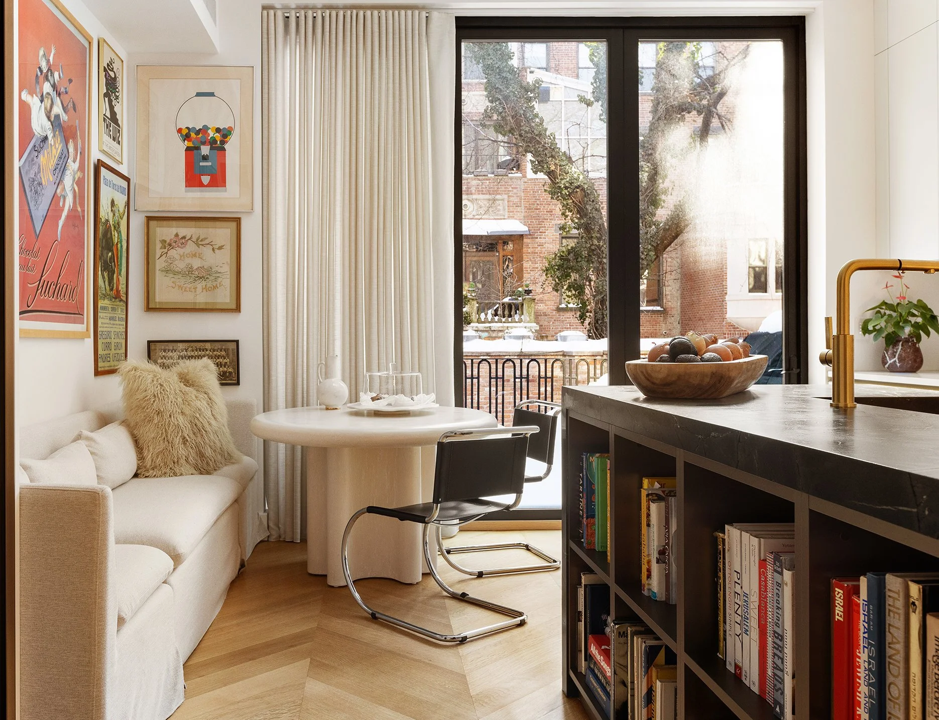 A cozy living room with a cream sofa, decorative throw pillows including a fuzzy one, a round white dining table with black chairs, a black bookshelf filled with books, large sliding glass doors leading to a balcony with trees and brick buildings out