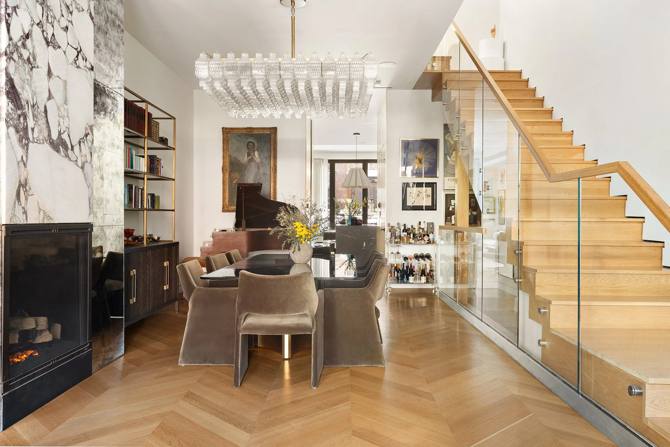 Modern living room with a dining area, featuring a large rectangular dining table with beige upholstered chairs, a fireplace with a marble surround, a chandelier, and a staircase with wooden steps and glass railing. The walls have framed artwork, and