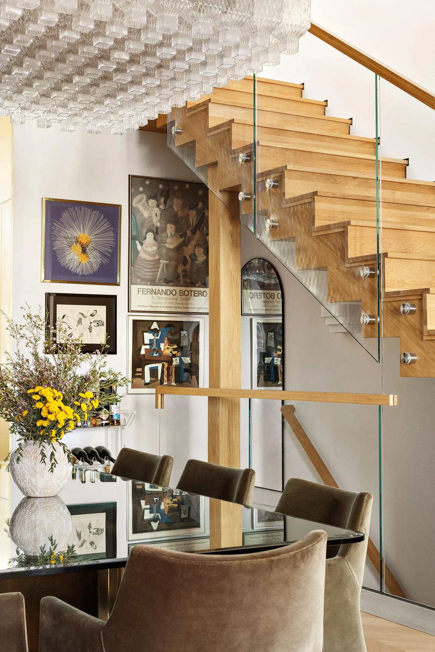 Interior of a modern home featuring a wooden staircase with glass railing, decorated with artwork on the wall, a large floral arrangement on a reflective dining table, and a contemporary glass chandelier overhead.