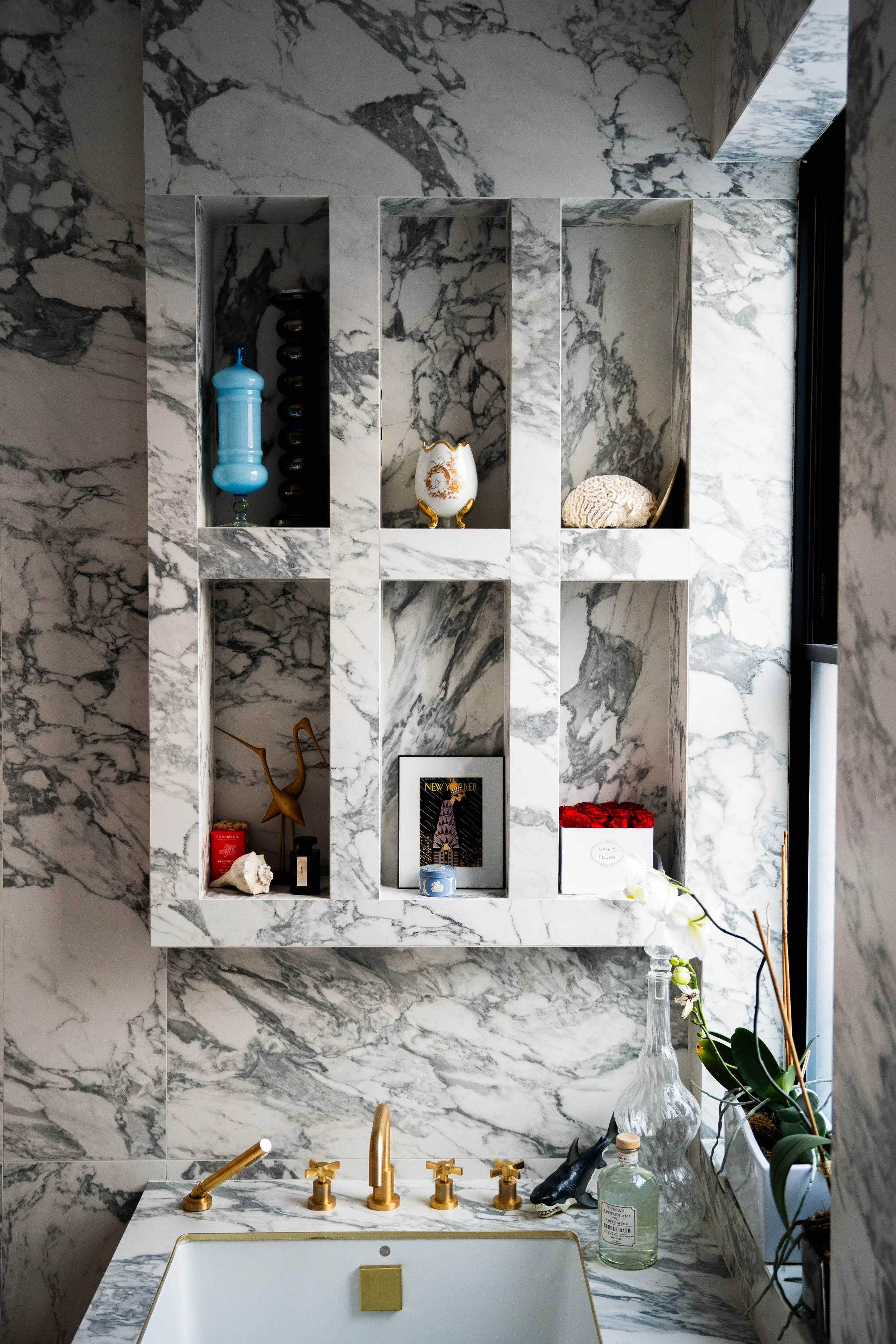 A marble bathroom with built-in shelves holding decorative items, a marble sink with gold fixtures, and a plant on the windowsill.