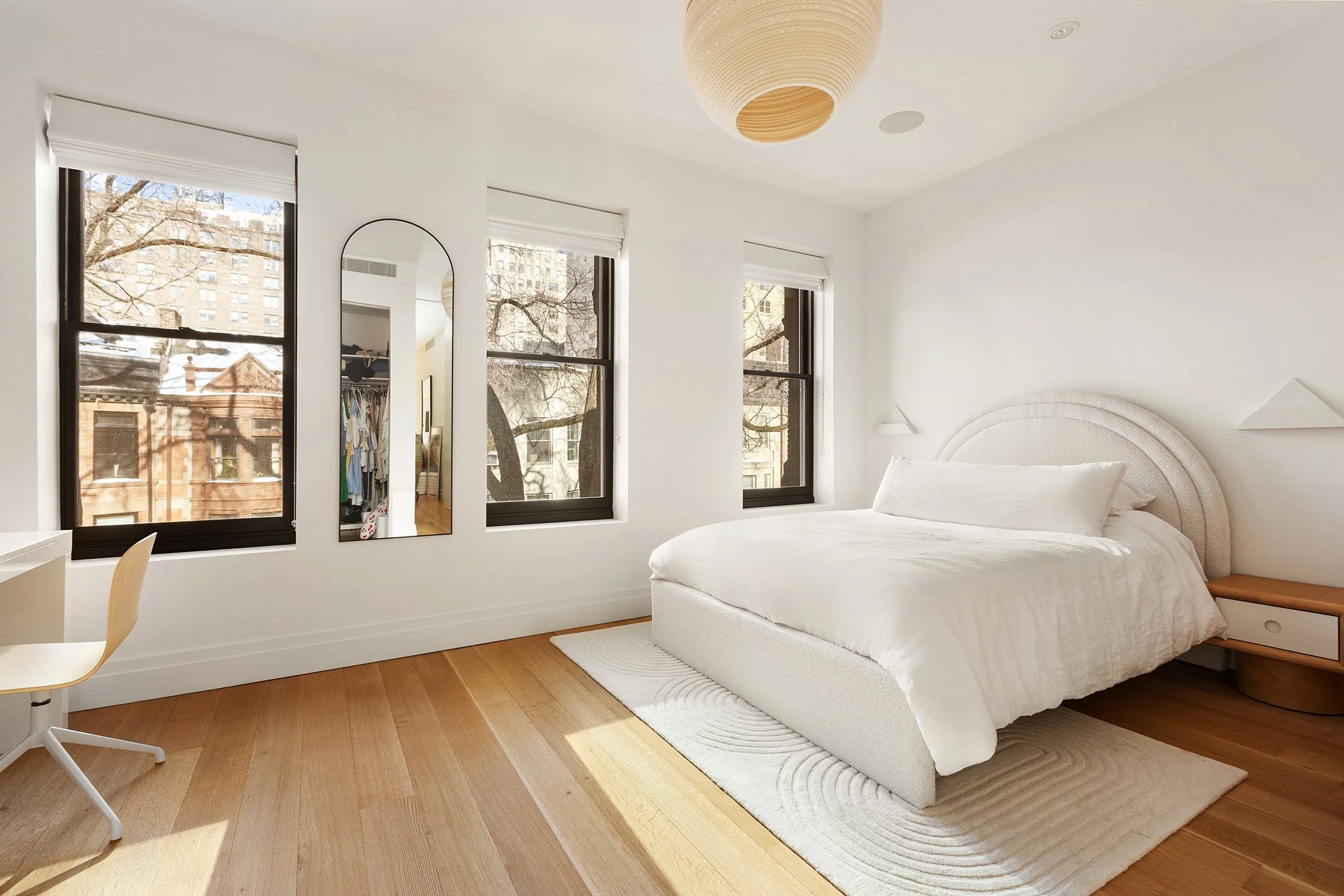 A bright bedroom with white walls, three windows showing a cityscape with trees and buildings, a white bed with a curved headboard, a wooden nightstand, a white rug, and a ceiling light fixture.