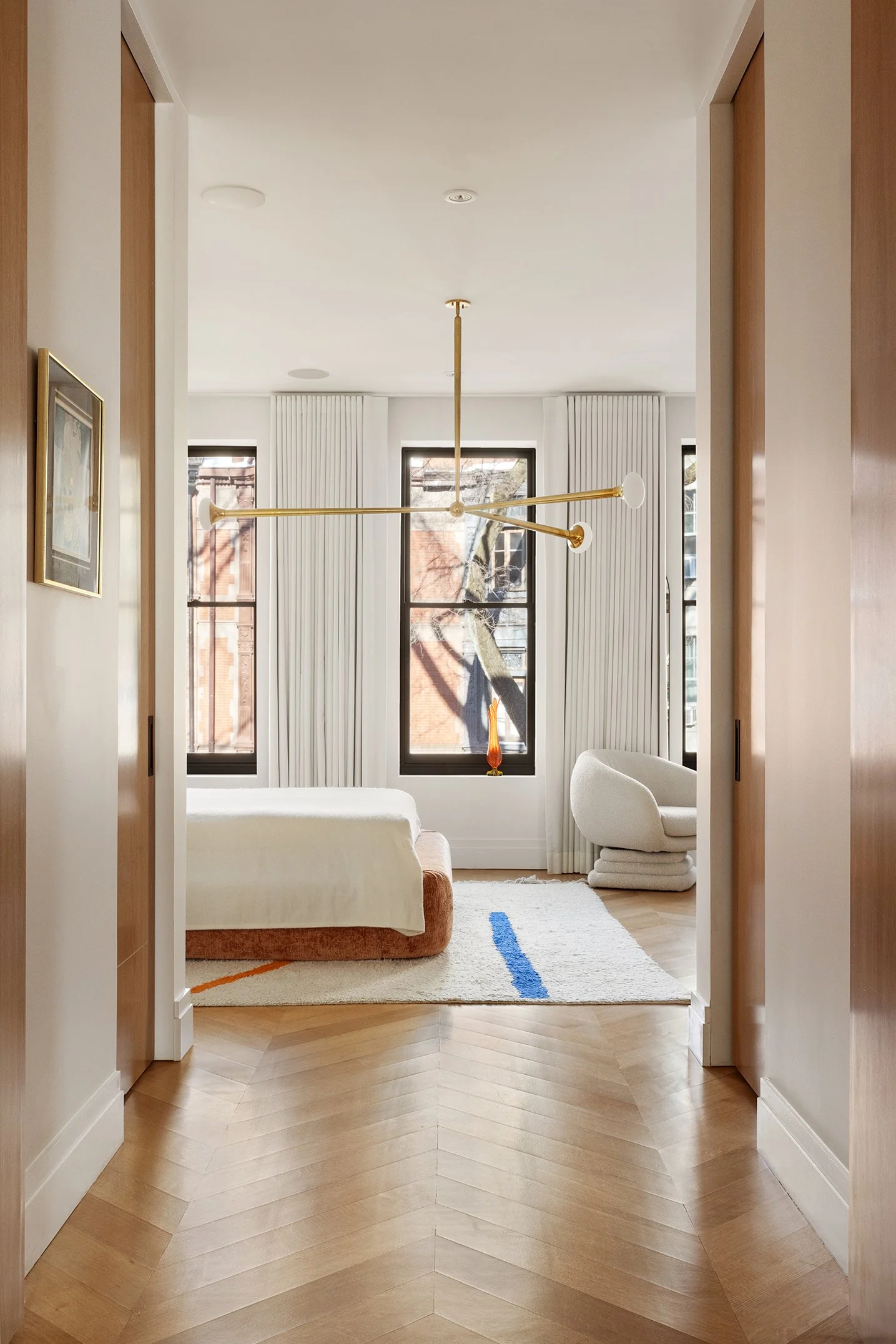 A modern bedroom with three large windows, white walls, light wood flooring, a white bed with a brown base, a white armchair, a white textured rug with colorful lines, and a gold pendant light fixture hanging from the ceiling.