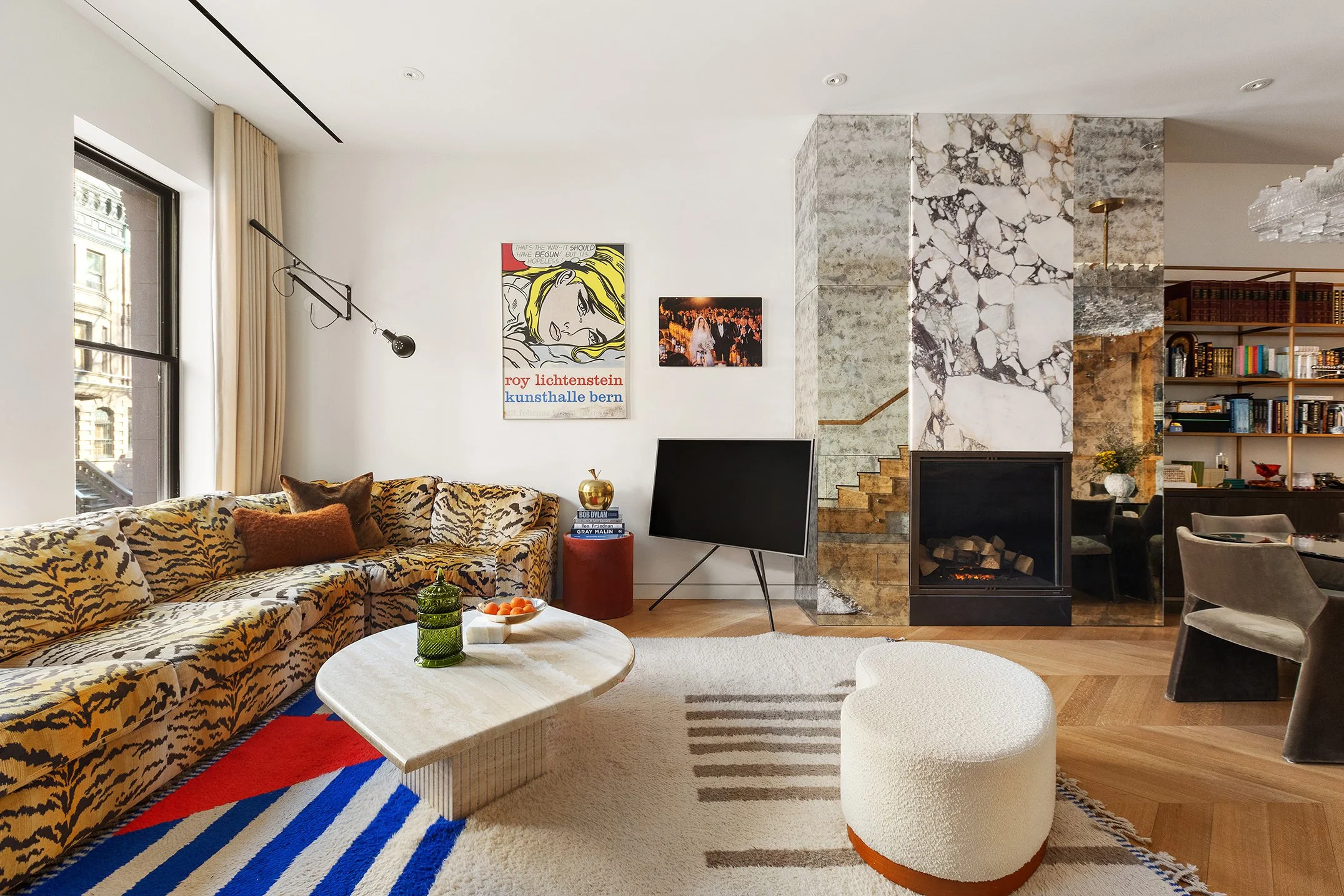 Living room with a large tiger-print sectional sofa, a white and blue striped rug, a cream ottoman, and a black TV on a stand. The walls are decorated with art, including a pop art piece and a photograph, with a marble fireplace and staircase in the 
