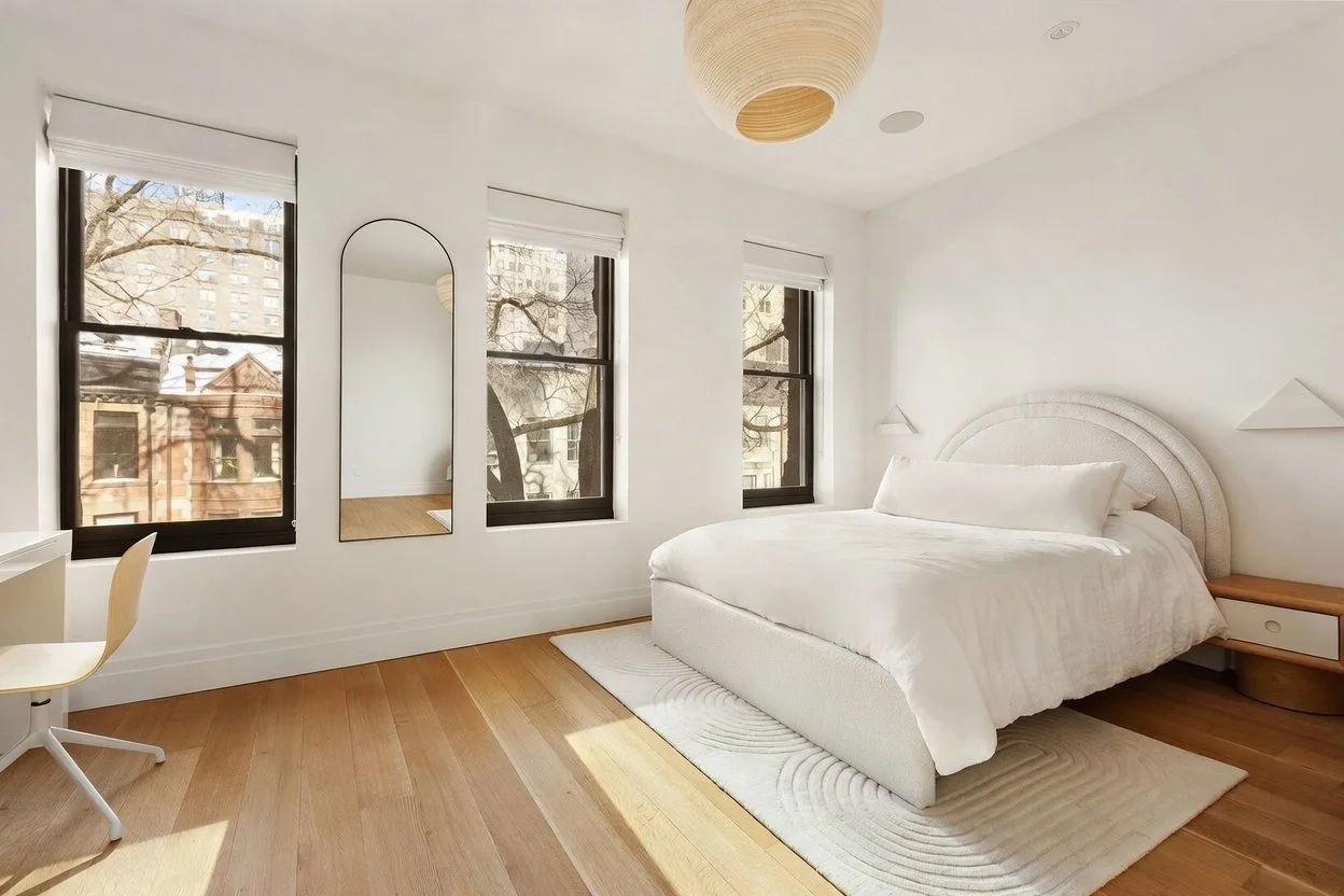 A bright, modern bedroom with three large windows, a white bed with an arched headboard and matching bedding, a white desk with a white chair, a round hanging light fixture, a full-length mirror, a small wooden nightstand, and a light-colored rug on 