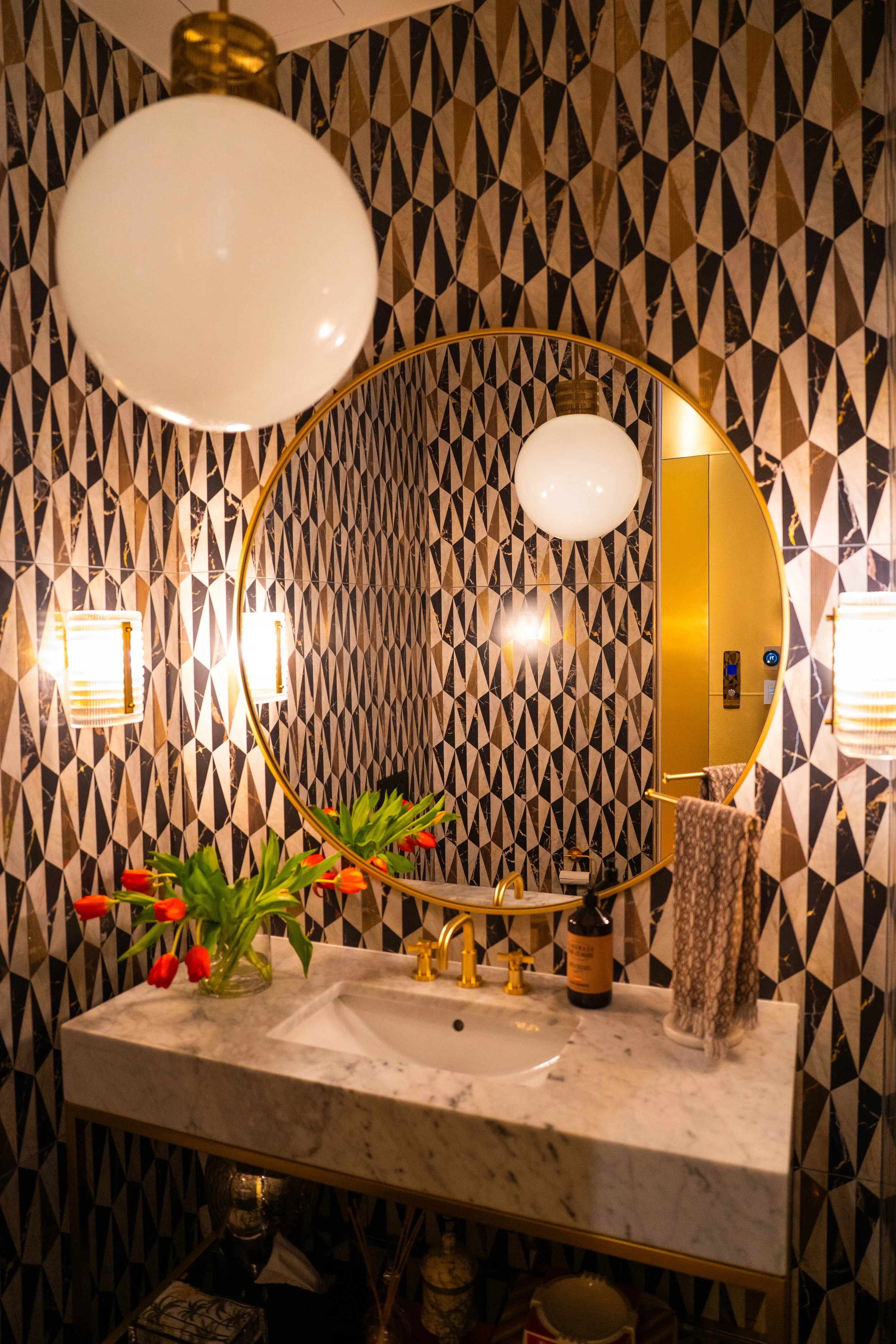 Decorative bathroom with patterned black and white wallpaper, round mirror, marble countertop, gold fixtures, vase of red tulips, soap bottle, and wall-mounted lights.