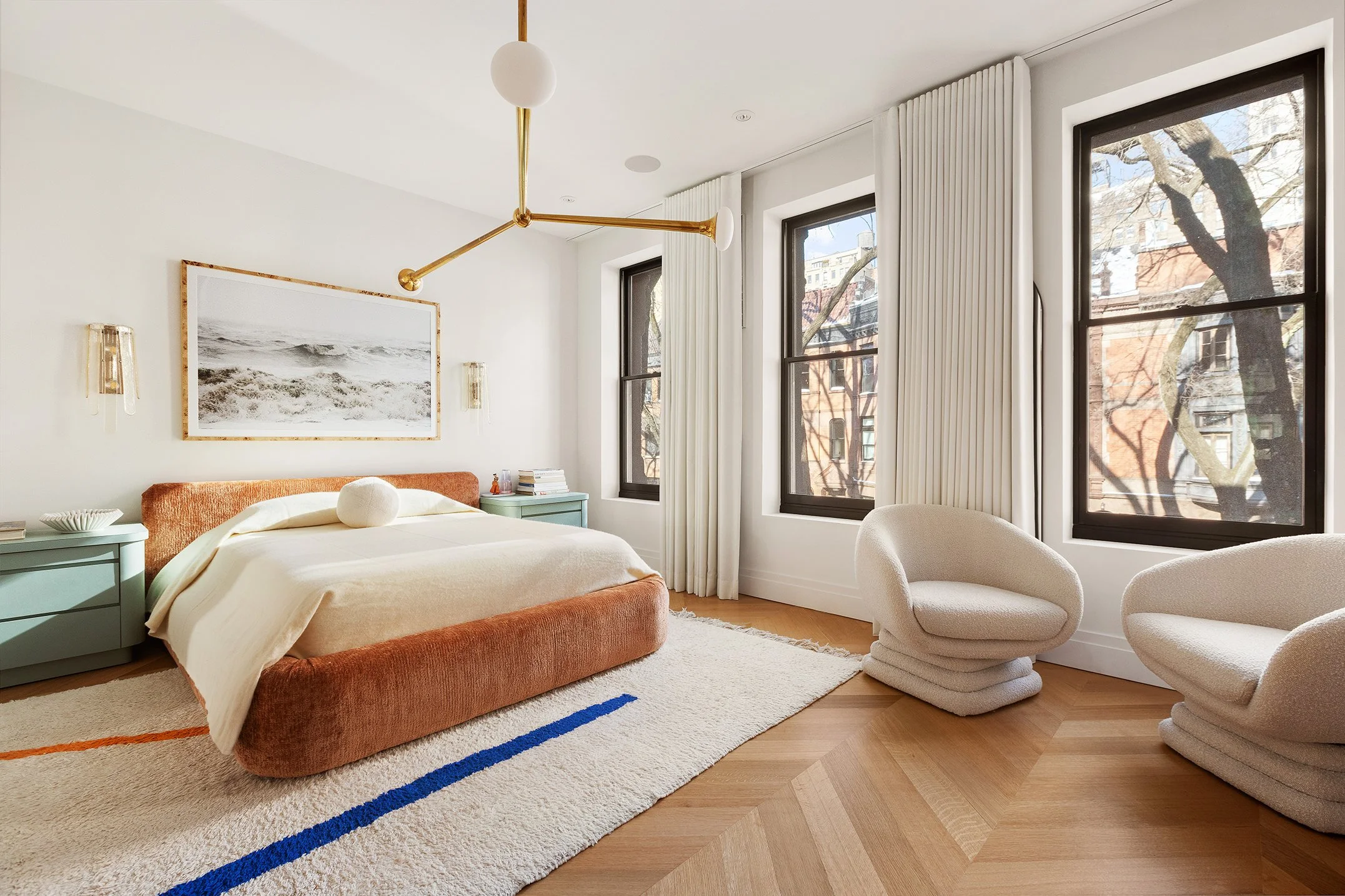 Modern bedroom with large windows, white curtains, and a plush bed with an orange velvet headboard. There are two pastel green nightstands, white chairs, and a contemporary gold and white chandelier. A framed black-and-white ocean photo is above the 