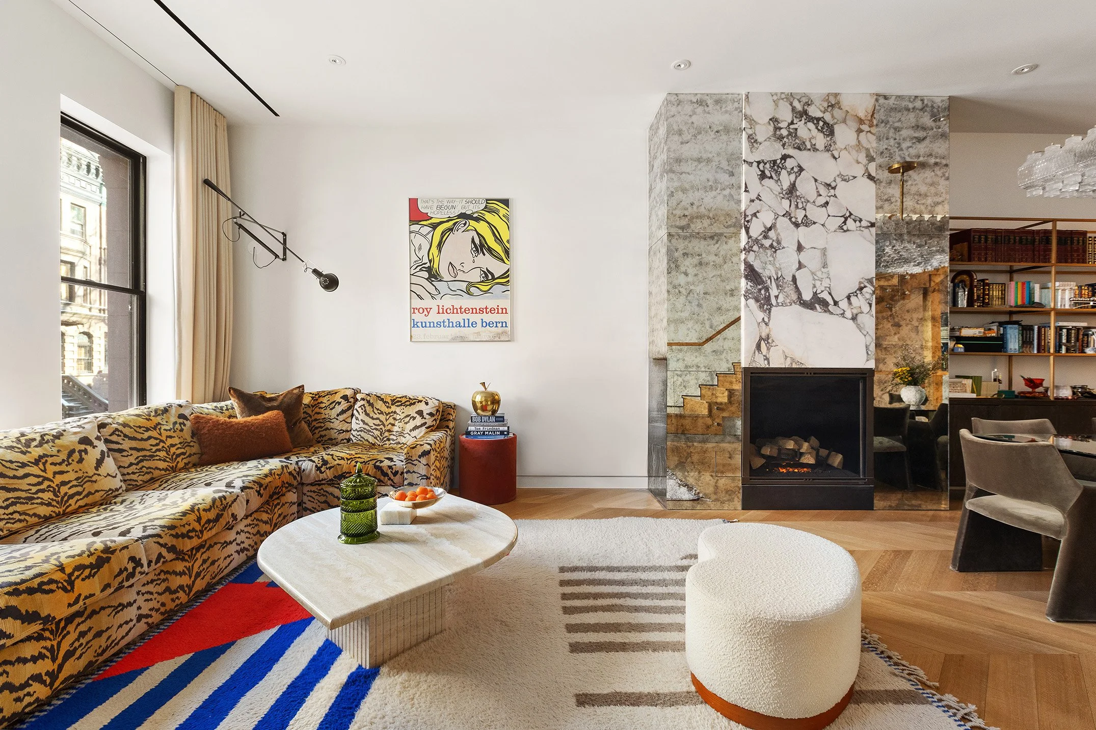 Living room with animal print sofa, round white ottoman, abstract art on white wall, fireplace, bookshelf, and staircase with marble accents.