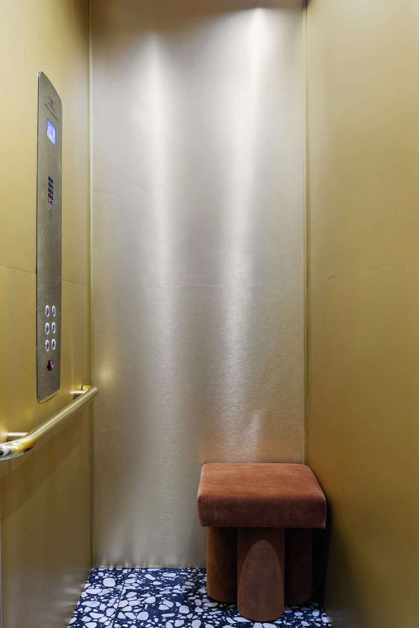 Interior of an elevator with a padded stool in the corner, gold walls, a silver door, and a control panel on the left wall.