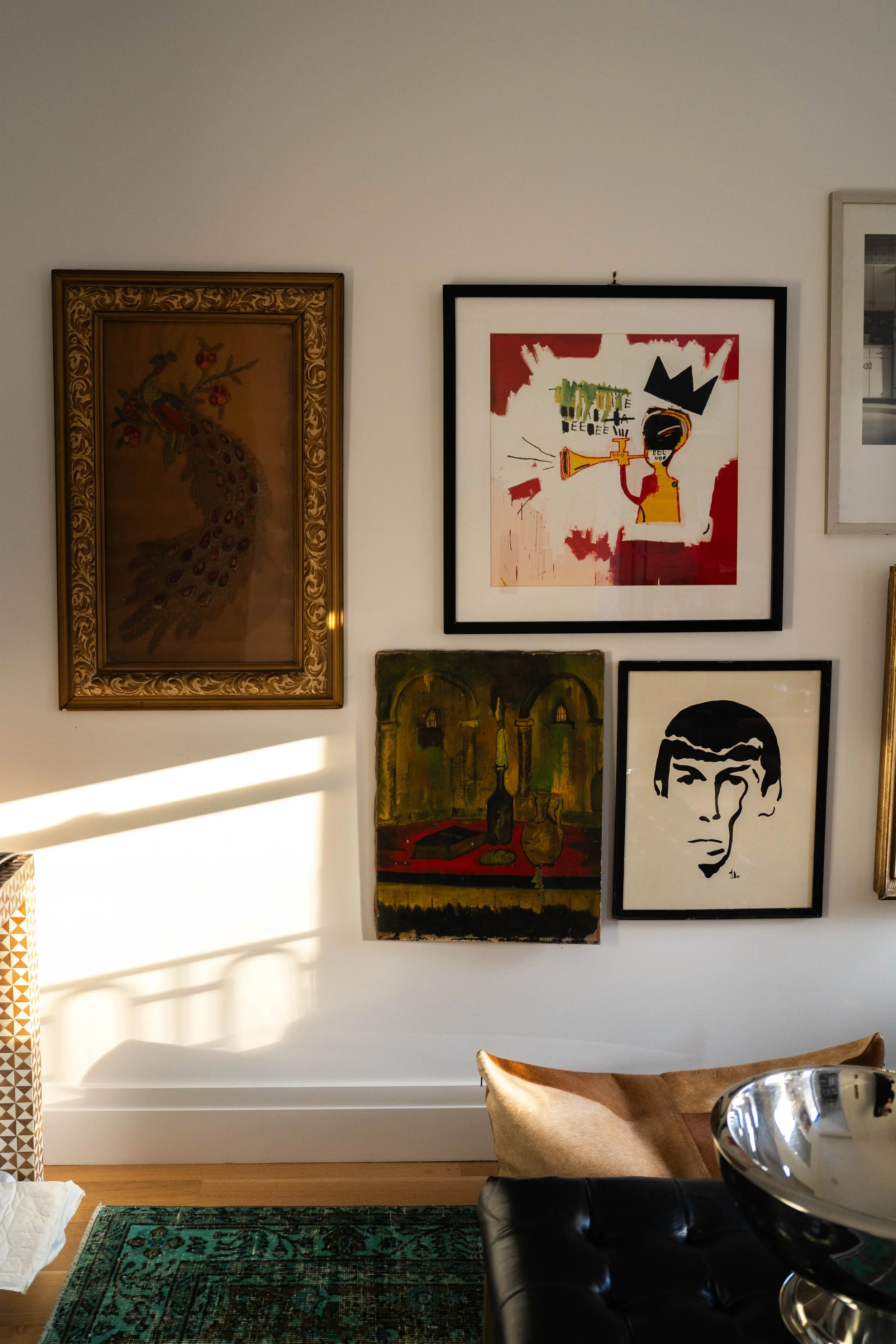 A wall with five framed artworks, including a peacock, an abstract figure with a crown, a still life with bottles, and a stylized portrait, in a room with a green rug and a partially visible leather chair.