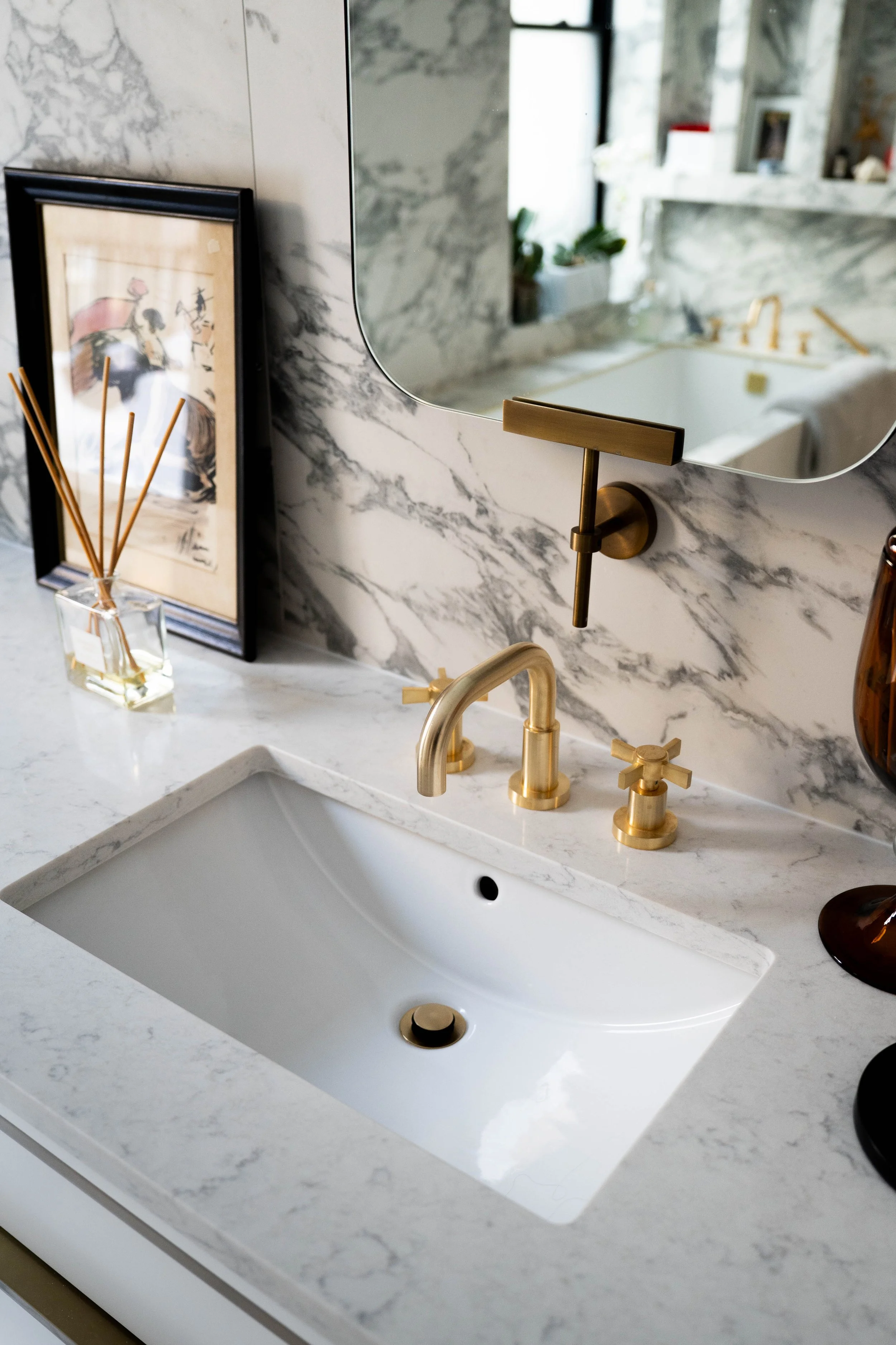 Bathroom vanity with marble countertop, white sink, gold faucet and handles, mirror with black trim, framed artwork, reed diffuser, and a brown vase.
