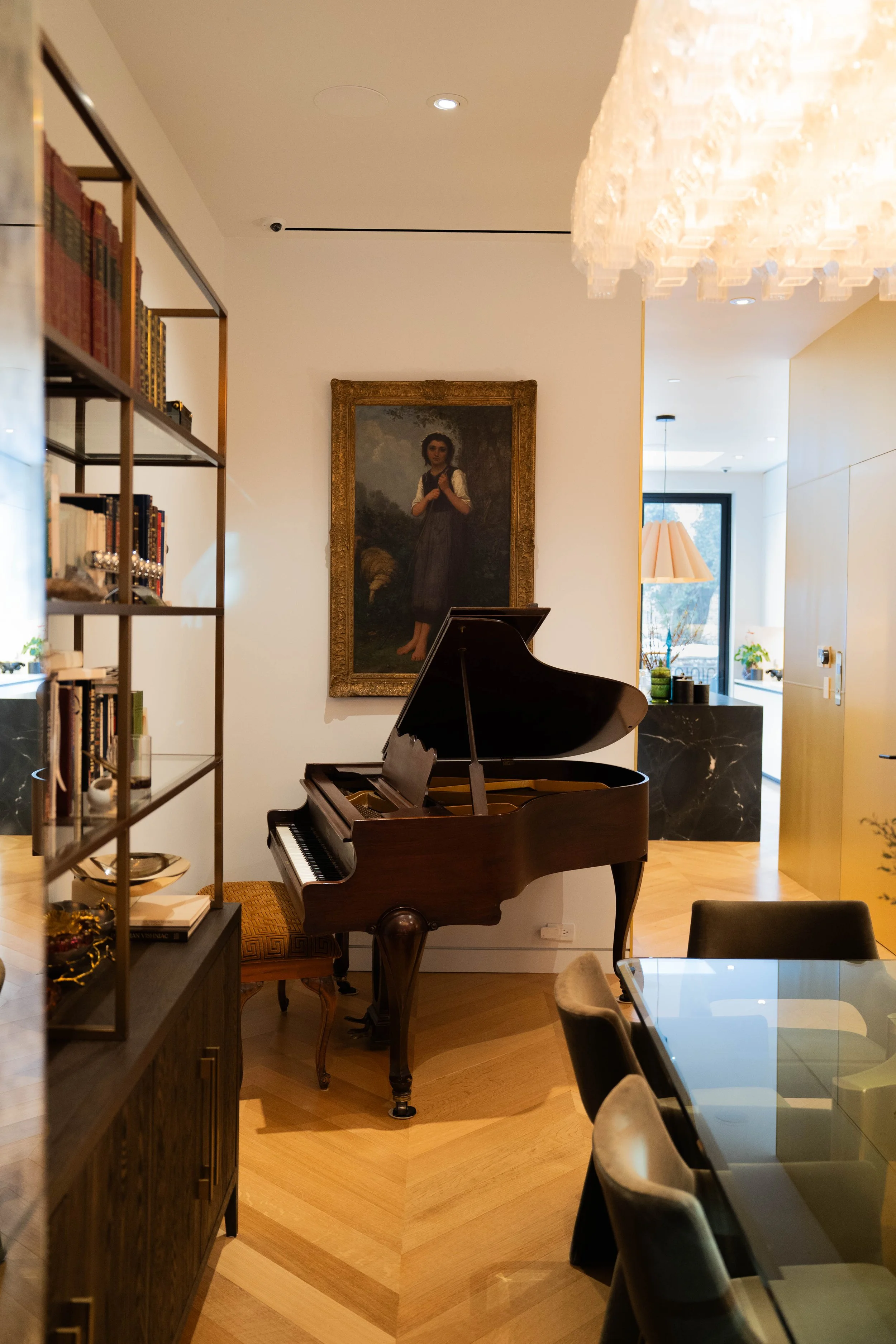 Interior of a living or dining area with a grand piano, a framed portrait hanging on the wall, a glass dining table with chairs, and a bookshelf with various books and decor items.