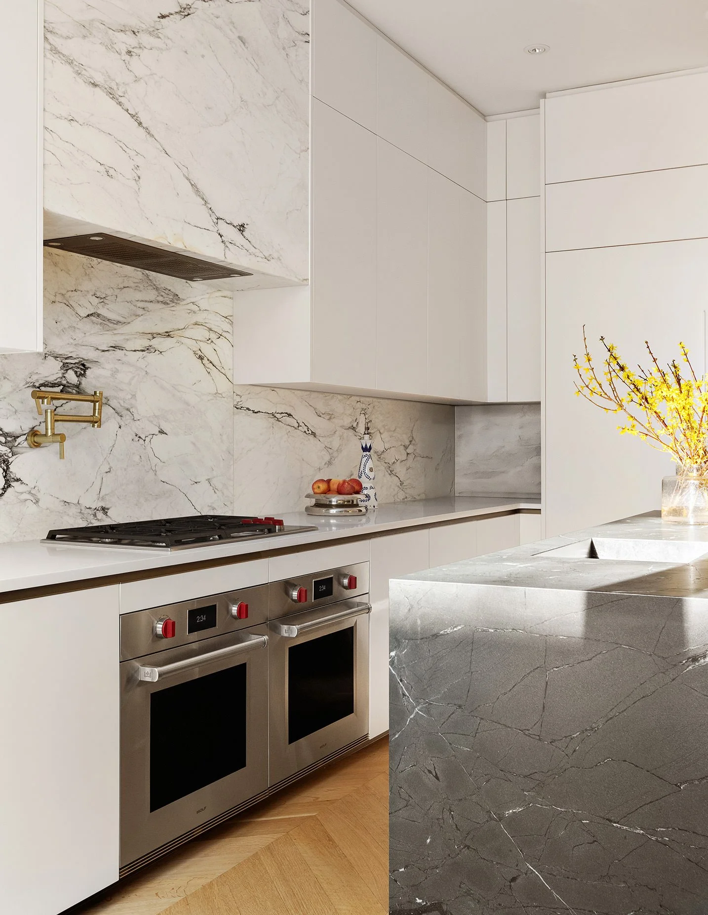 Modern kitchen with white cabinetry, marble backsplash, stainless steel ovens, and a marble countertop island with a vase of yellow flowers.