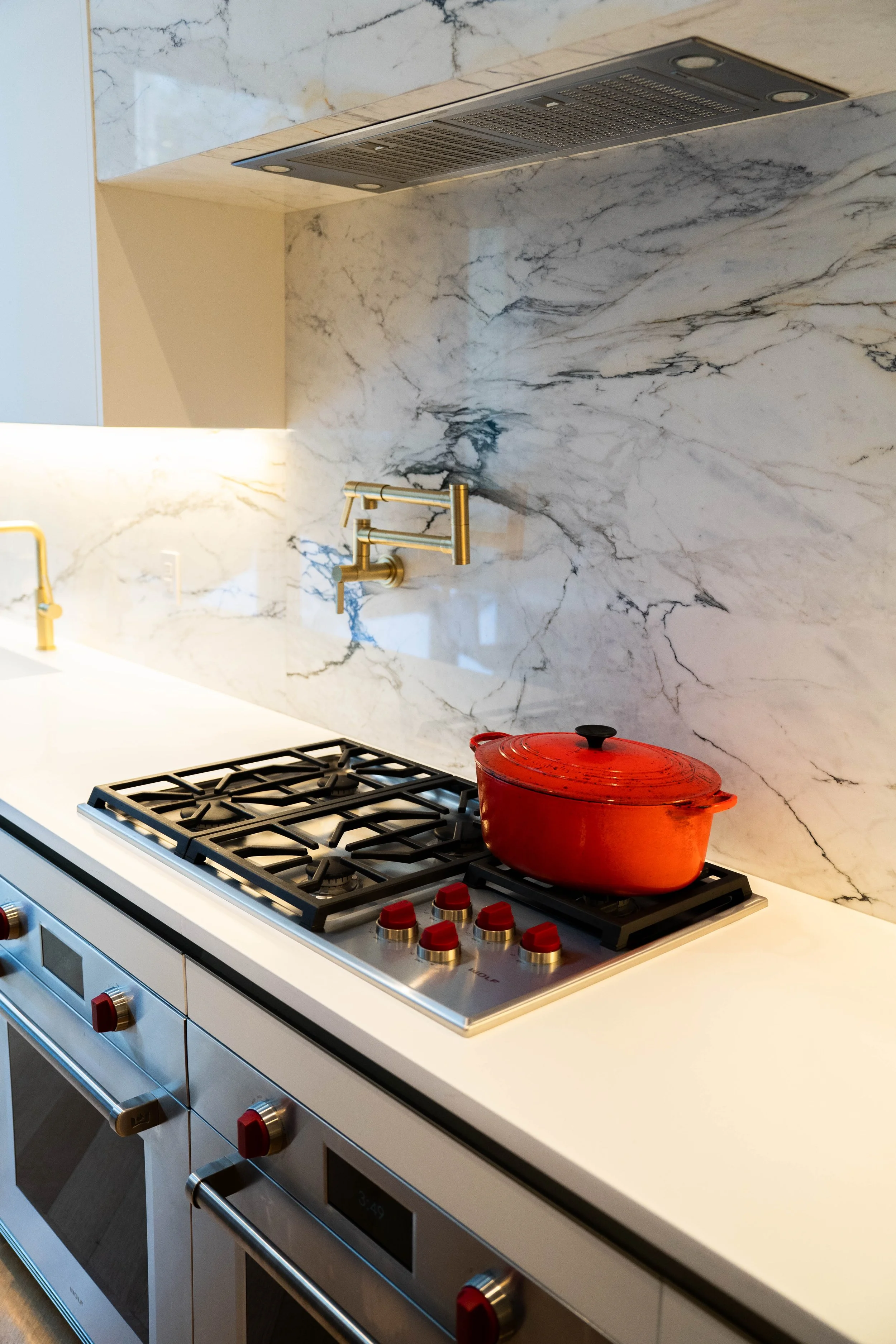 Modern kitchen with a white countertop, a red cooking pot on a gas stove, and a marble backsplash with gold fixtures and accents.