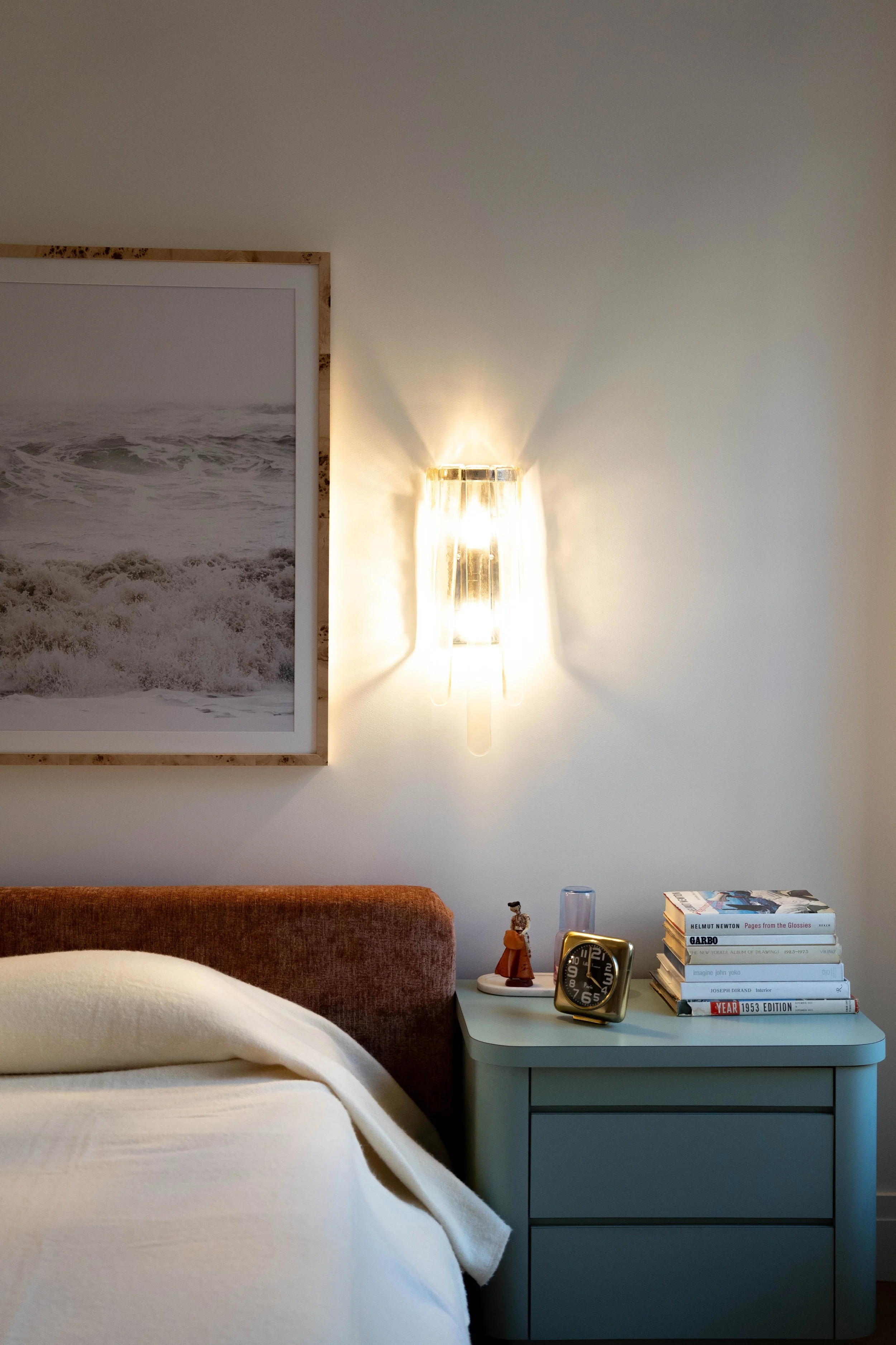 A bedroom with a nightstand, a lamp, a stack of books, a framed wave photograph, and a bed with cream-colored bedding.