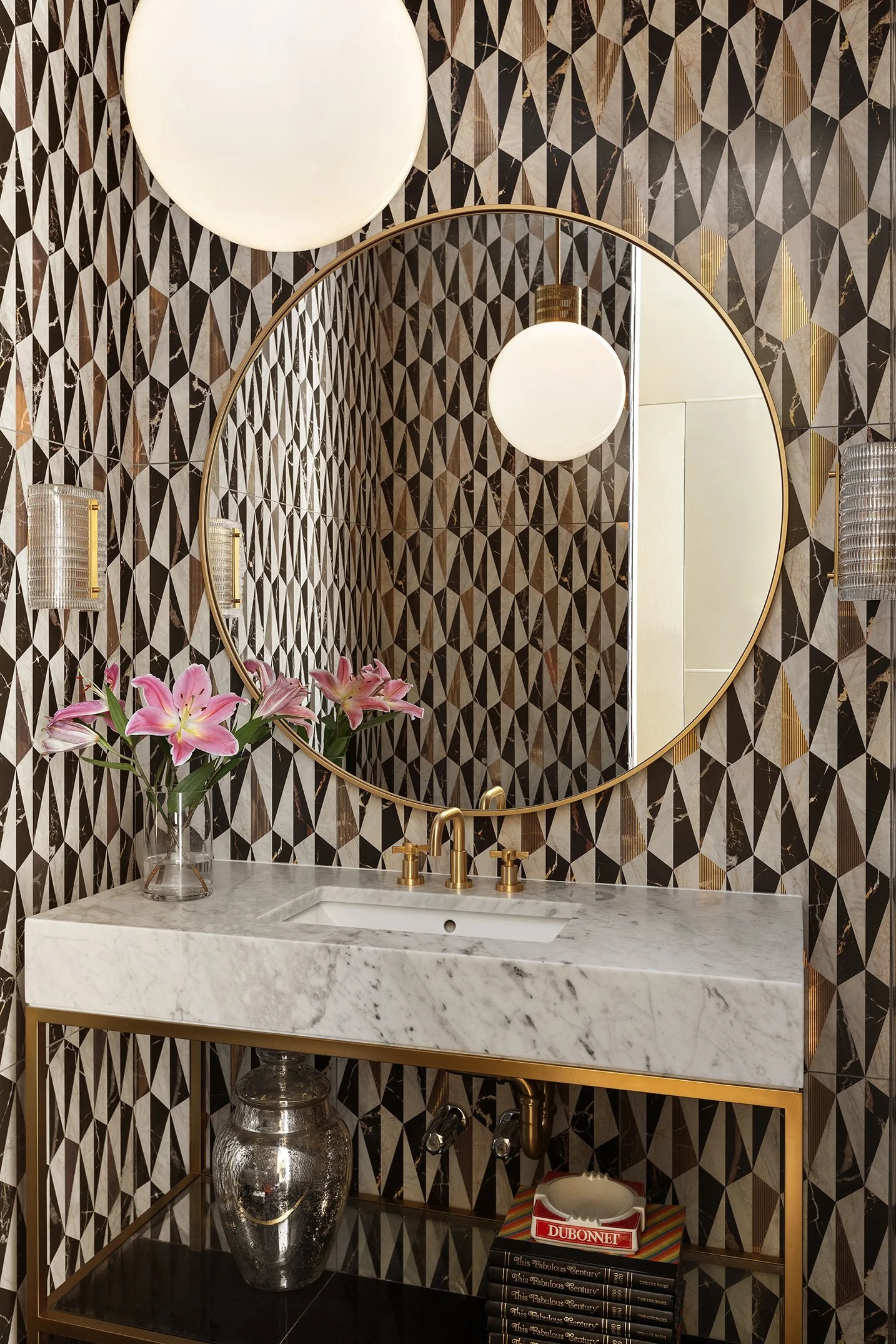 Bathroom with geometric patterned wallpaper, round mirror, white marble sink with brass hardware, pink lilies in a vase, and a wall-mounted light fixture.