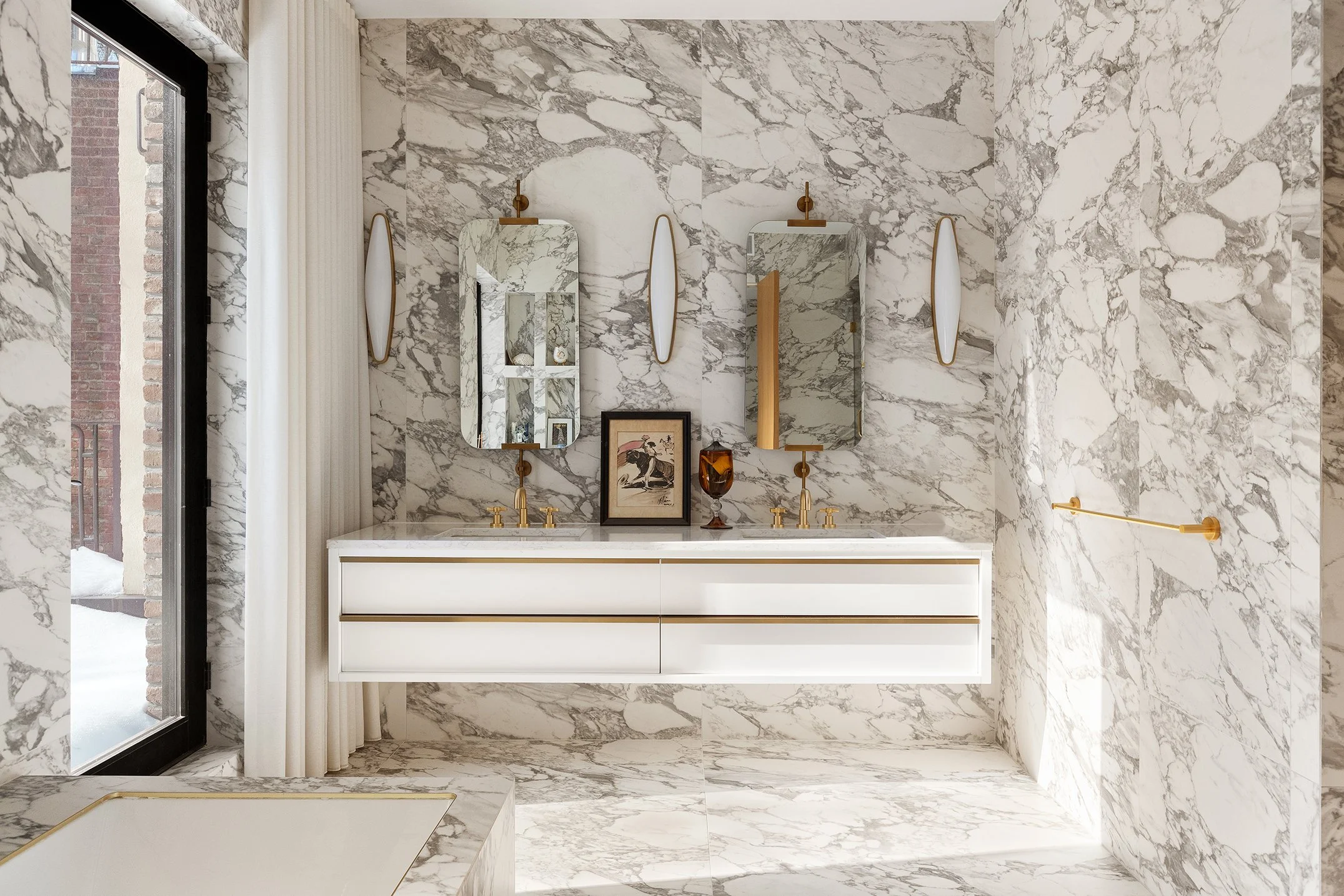 A luxurious bathroom with marble walls, a white floating double vanity with gold accents, and two mirrors above each sink. There are two vertical wall sconces and a framed artwork between the mirrors. A sliding glass door with sheer white curtains le