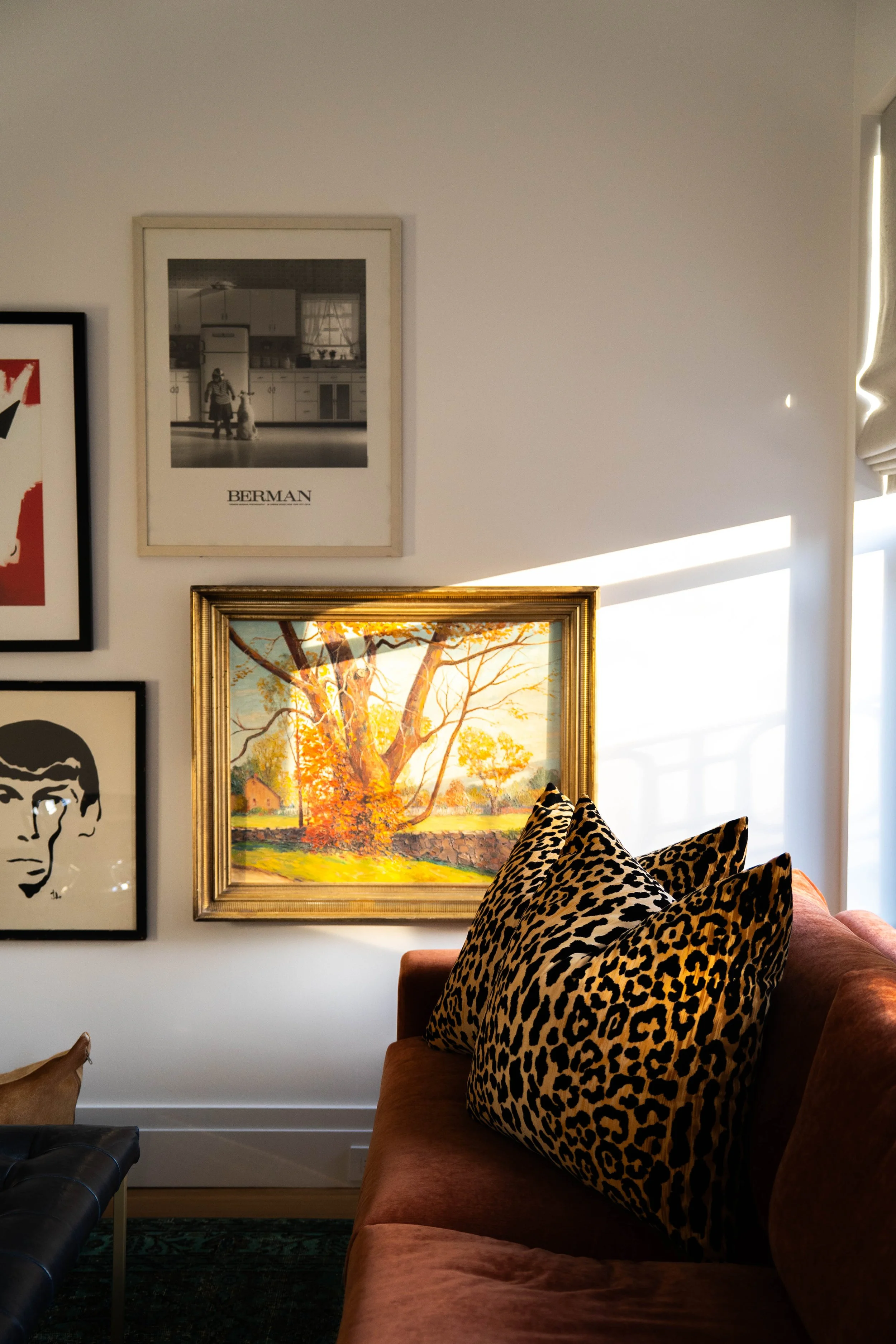 Living room wall with framed art and paintings, including a black and white photo labeled 'BERMAN,' a colorful landscape, and artwork of a person's face, with a sofa showing leopard print pillows and sunlight streaming in through a window.