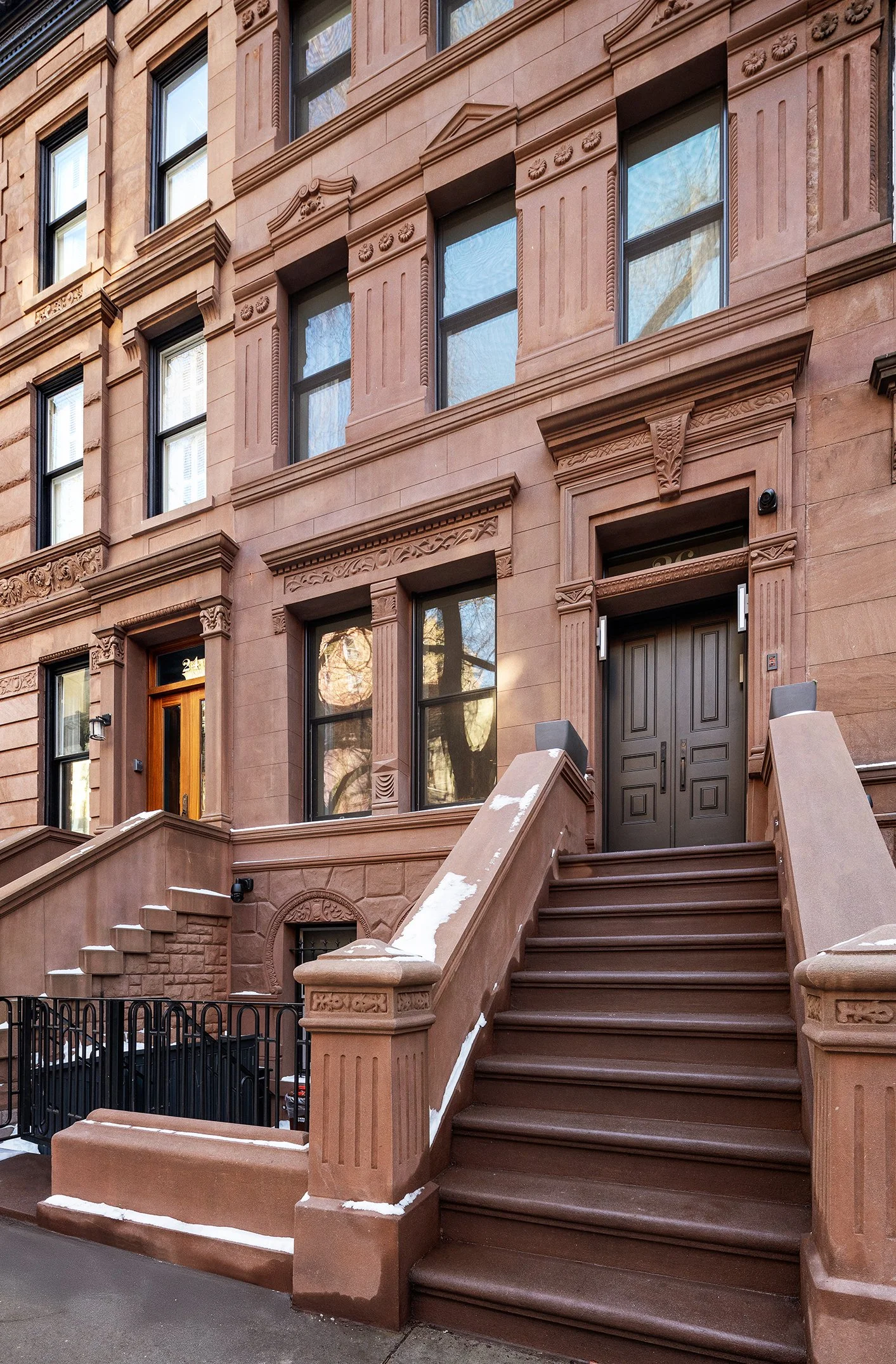 Brownstone apartment building with stairs leading up to dark-colored front doors, multiple windows, decorative architectural details, and some snow on the stairs.