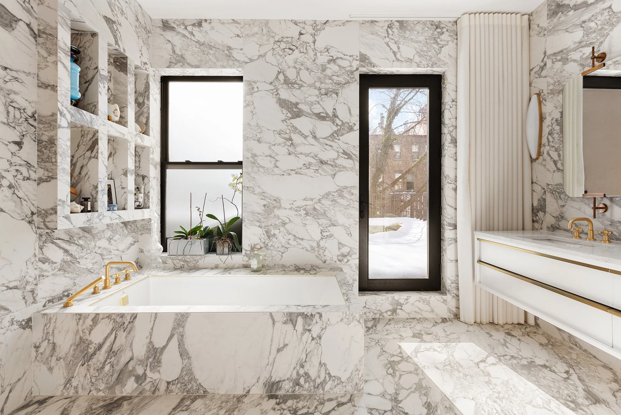 Luxurious bathroom with white marble walls and floor, featuring a built-in bathtub with gold fixtures, two black-framed windows, a white vanity with gold hardware, and a wall-mounted mirror.
