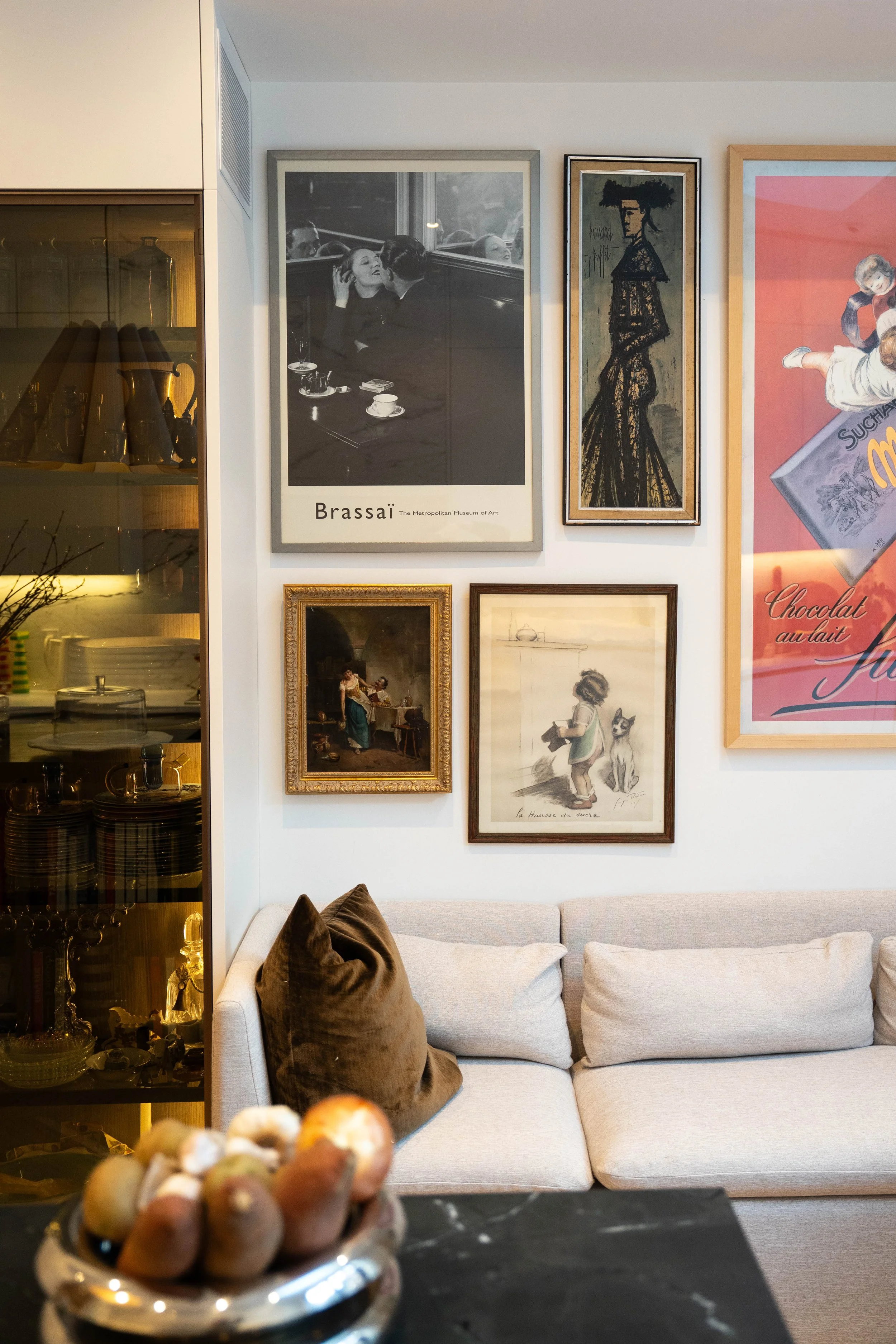 Living room with a beige sofa, framed artwork on the wall, a dark coffee table with bread rolls, and a glass cabinet with dishes on the left side.