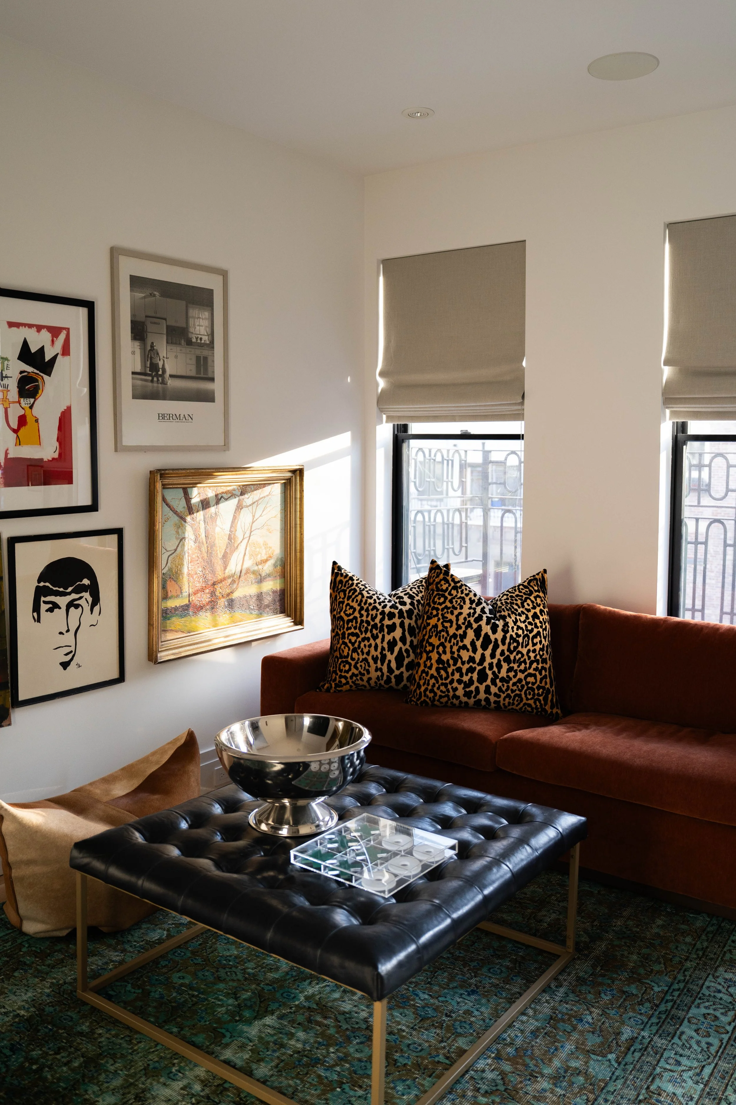 A living room with a black tufted coffee table, a brown sofa with leopard print pillows, and various framed artwork on the white wall. Two windows with beige blinds let in natural light.