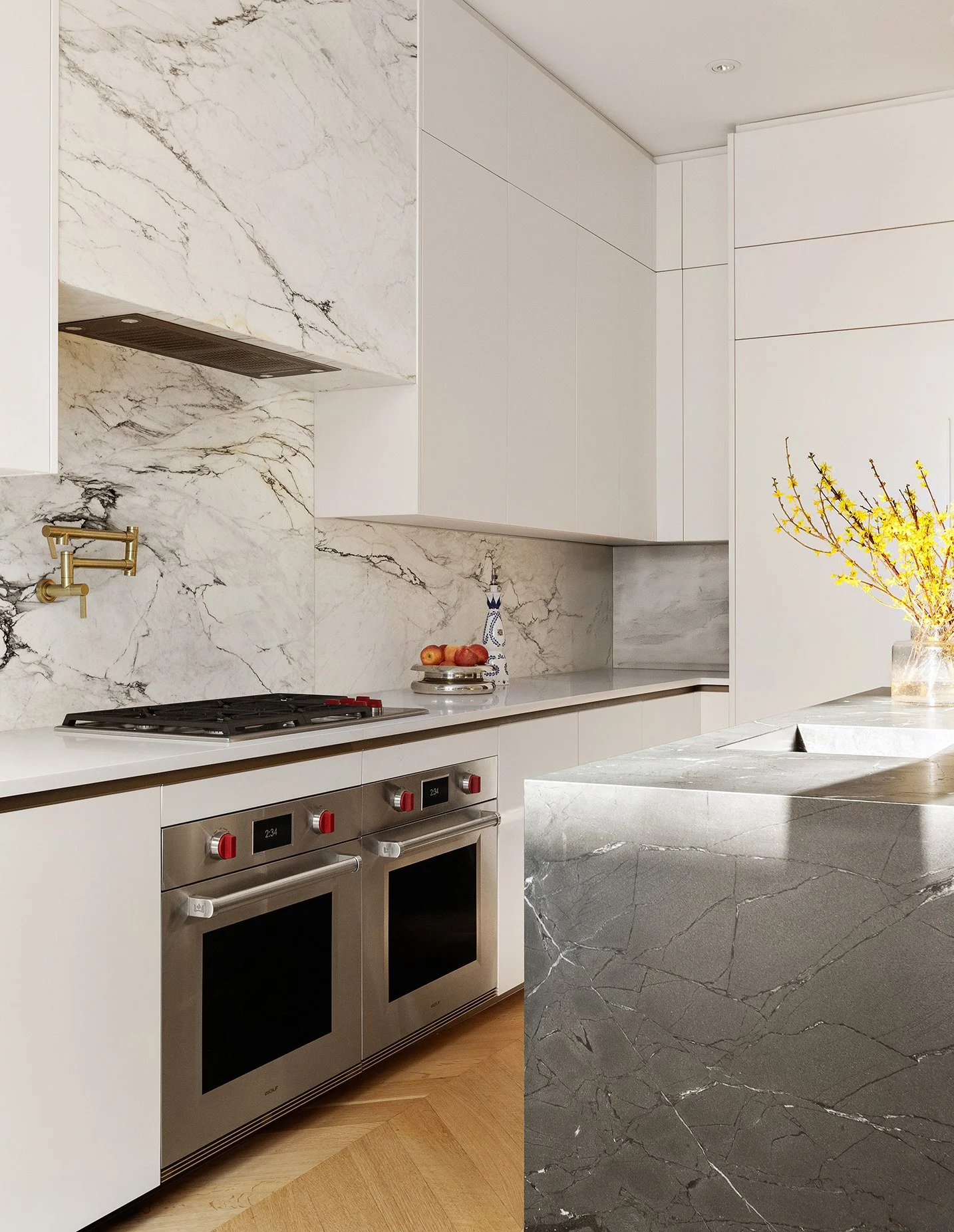 Modern kitchen with white cabinets, marble backsplash, and a marble kitchen island. Stainless steel oven and stove with red control knobs, a brass faucet on the wall, and a vase with yellow flowers on the island.