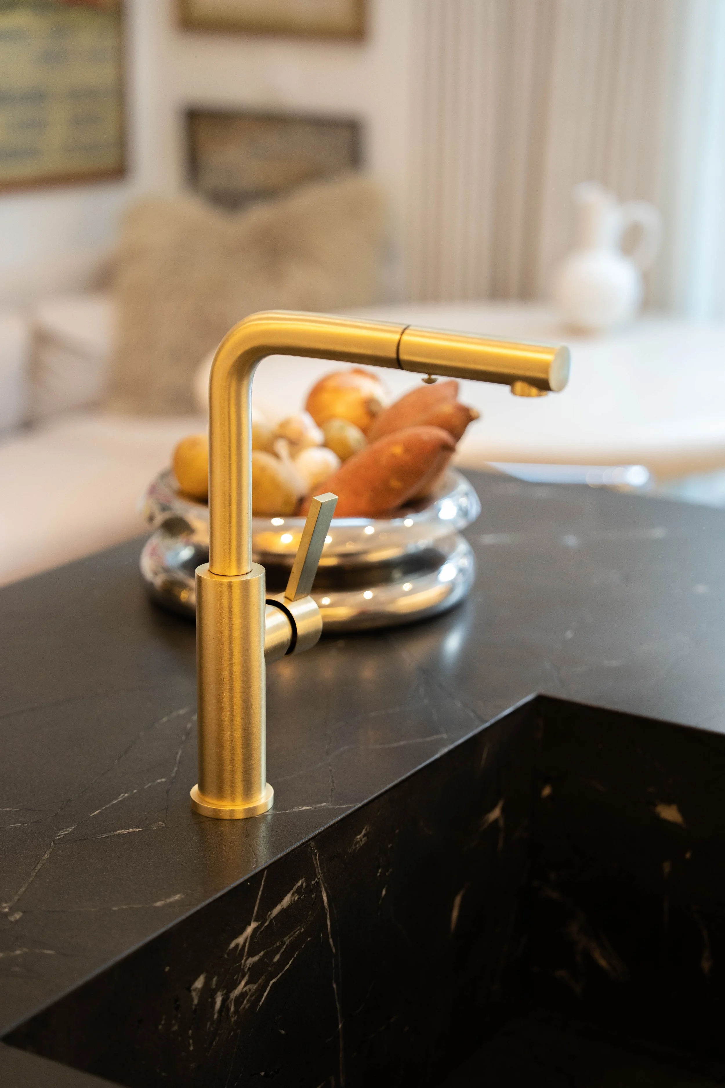 Close-up of a gold-colored kitchen sink faucet on a black marble countertop, with a stainless steel bowl of fruit in the background.