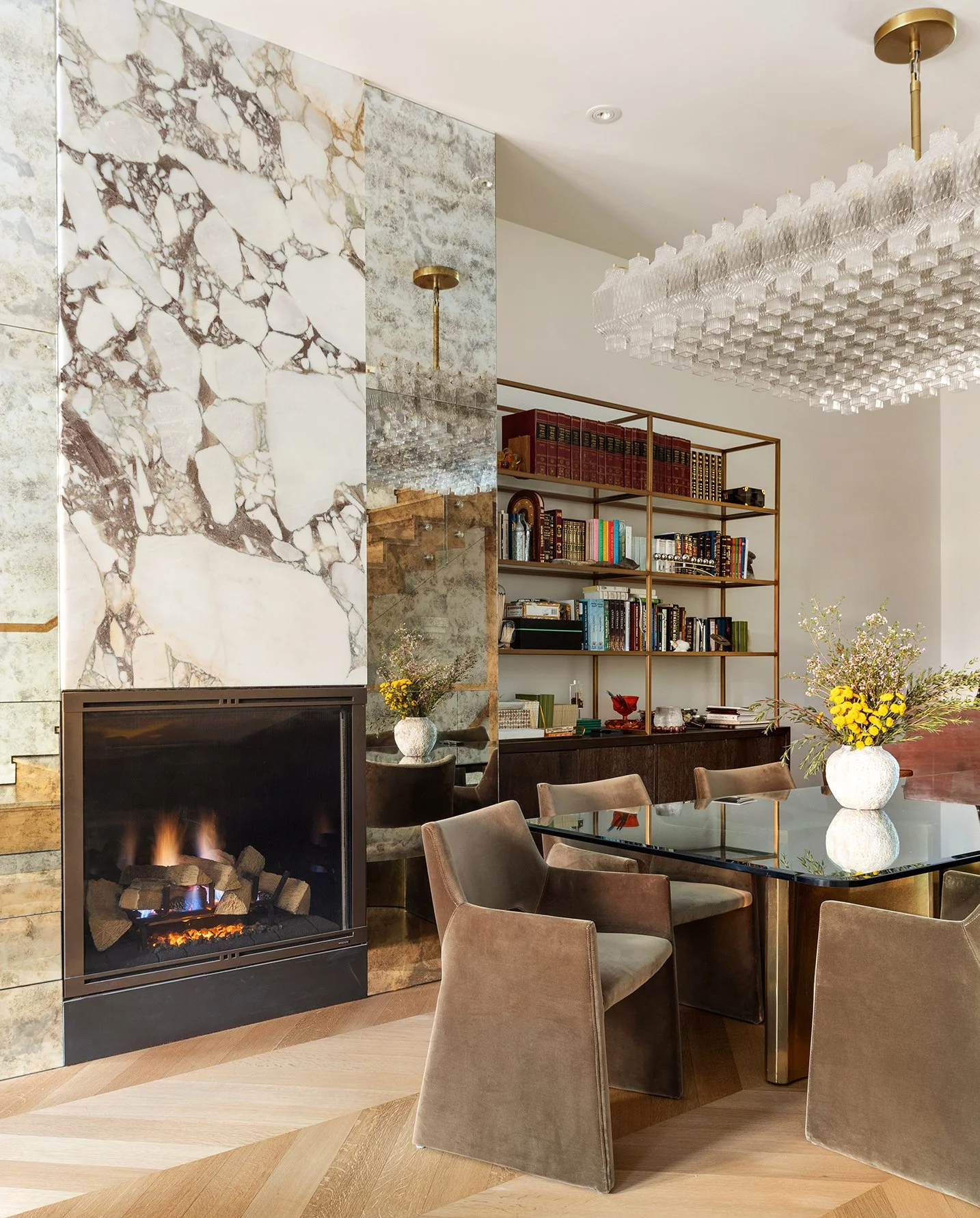 A contemporary living room features a marble and stone fireplace, a glass-top dining table with beige upholstered chairs, a modern chandelier, a bookshelf with books and decorative items, and floral arrangements in vases.