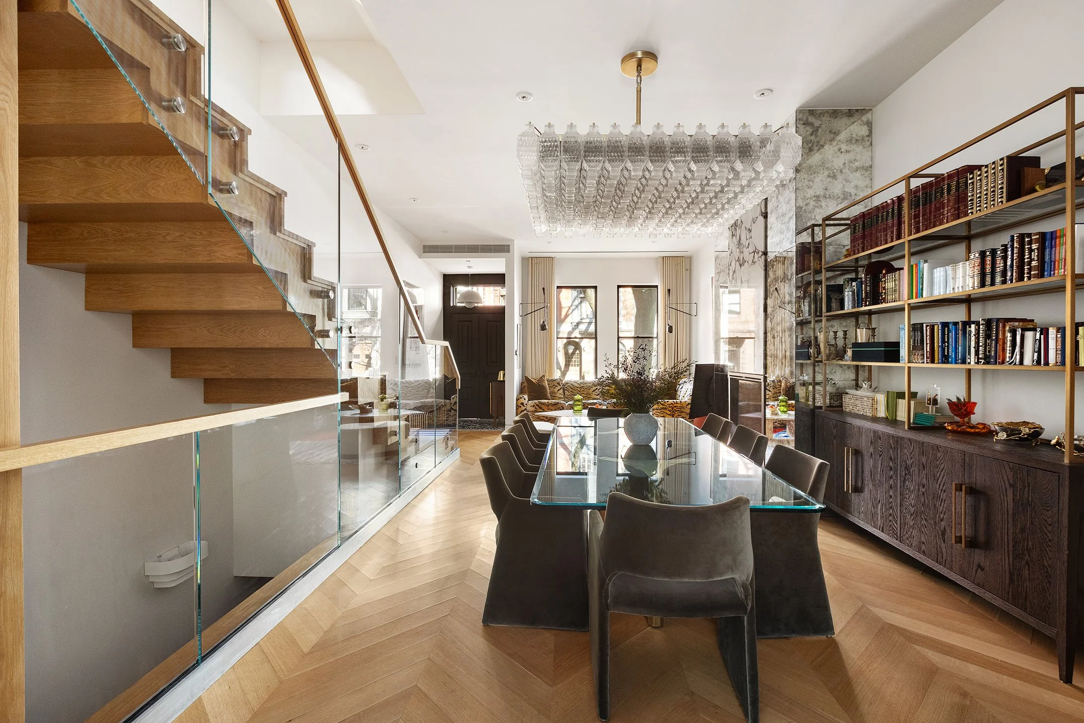 Modern living room with a glass dining table, gray chairs, bookshelf, staircase with glass railing, and large windows.