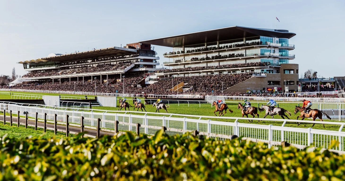 Horse race at a packed racetrack with a modern, multi-story grandstand filled with spectators.