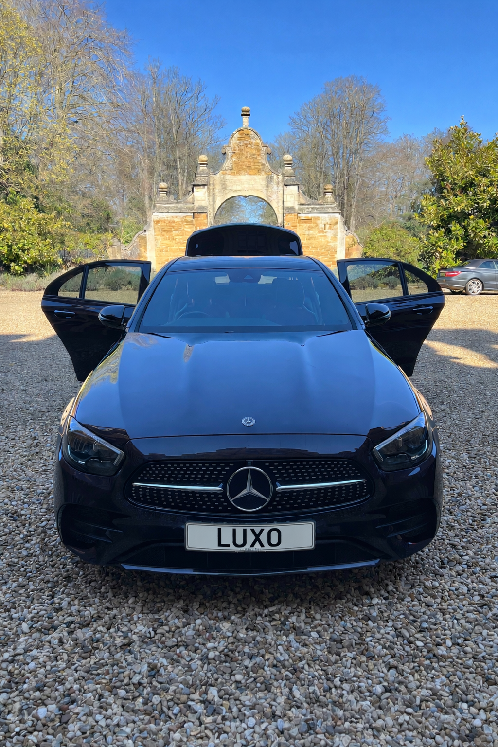 Black Mercedes-Benz car with open doors and a custom license plate that reads 'LUXO', parked on a gravel surface with an ornate stone archway and trees in the background.